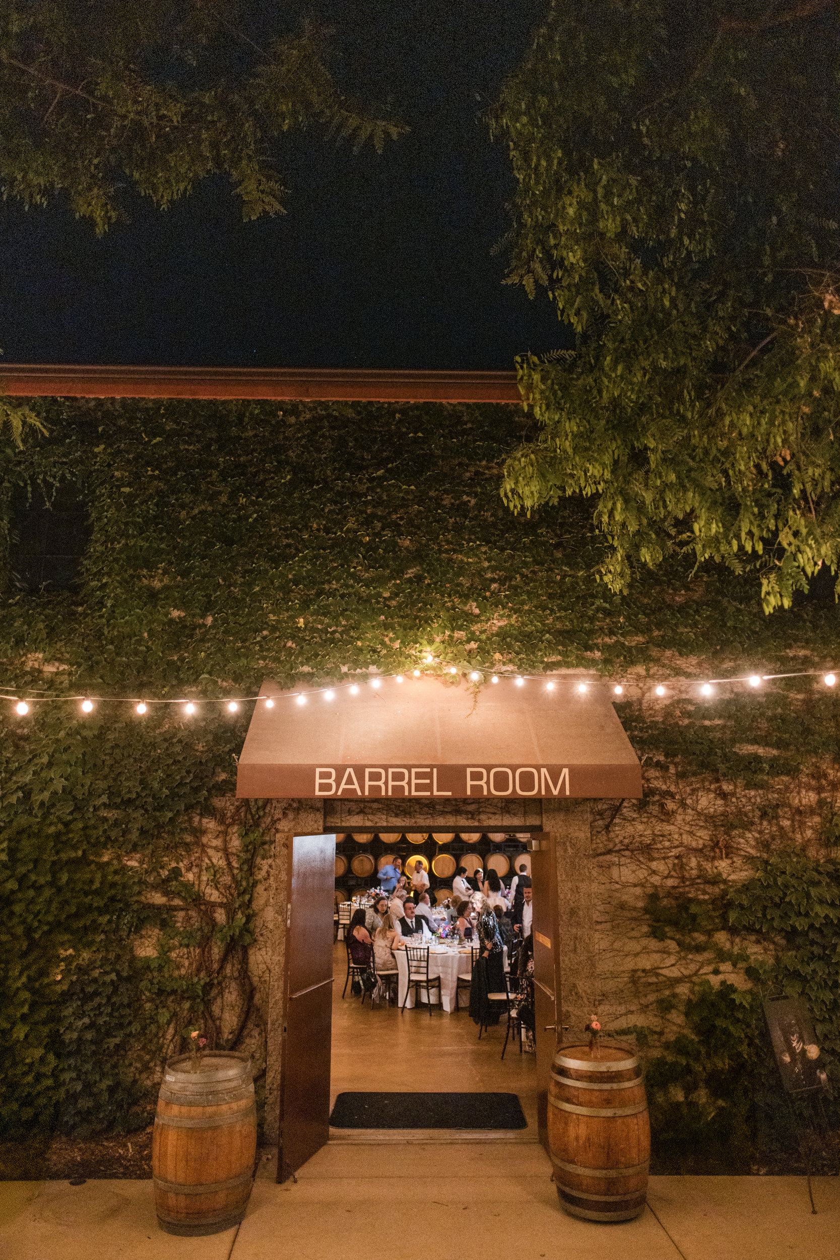 Exterior view of the Barrel Room at night, with people dining inside, string lights overhead, and two barrels by the entrance—an inviting scene perfect for a memorable Leoness Cellars wedding celebration.