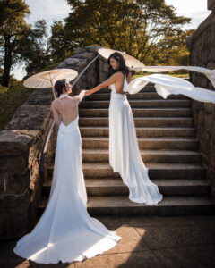 Two brides in white dresses stand on outdoor stone steps at a New York Botanical Garden wedding, holding parasols. One stands above, reaching out to the other, with trees and sunlight in the background.