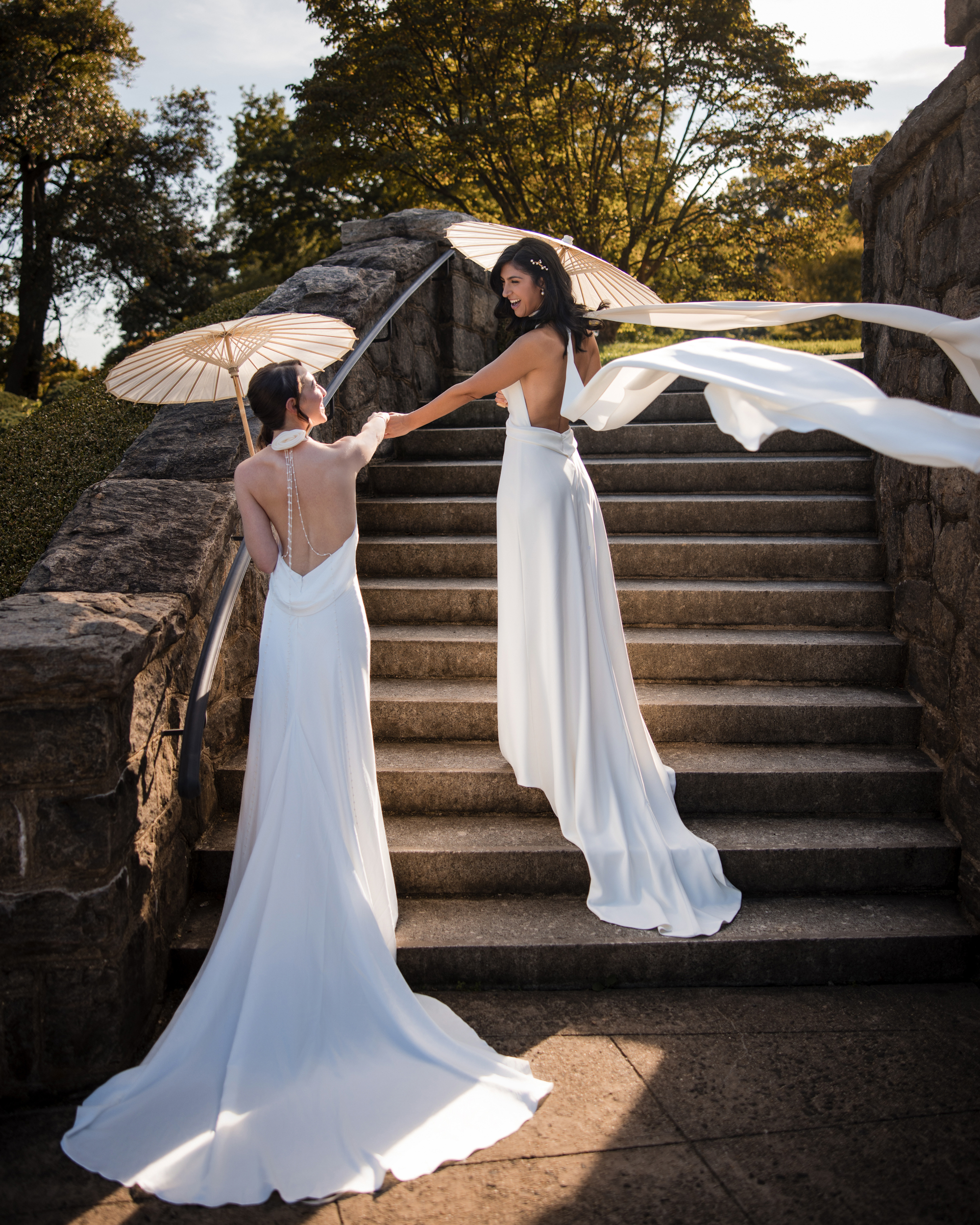 Two brides in white dresses stand on outdoor stone steps at a New York Botanical Garden wedding, holding parasols. One stands above, reaching out to the other, with trees and sunlight in the background.