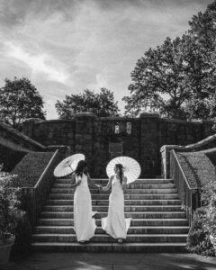 Two brides in white dresses holding parasols ascend outdoor stone steps surrounded by trees and shrubs, with a stone wall visible at the top—a timeless black and white photo evoking a New York Botanical Garden wedding.