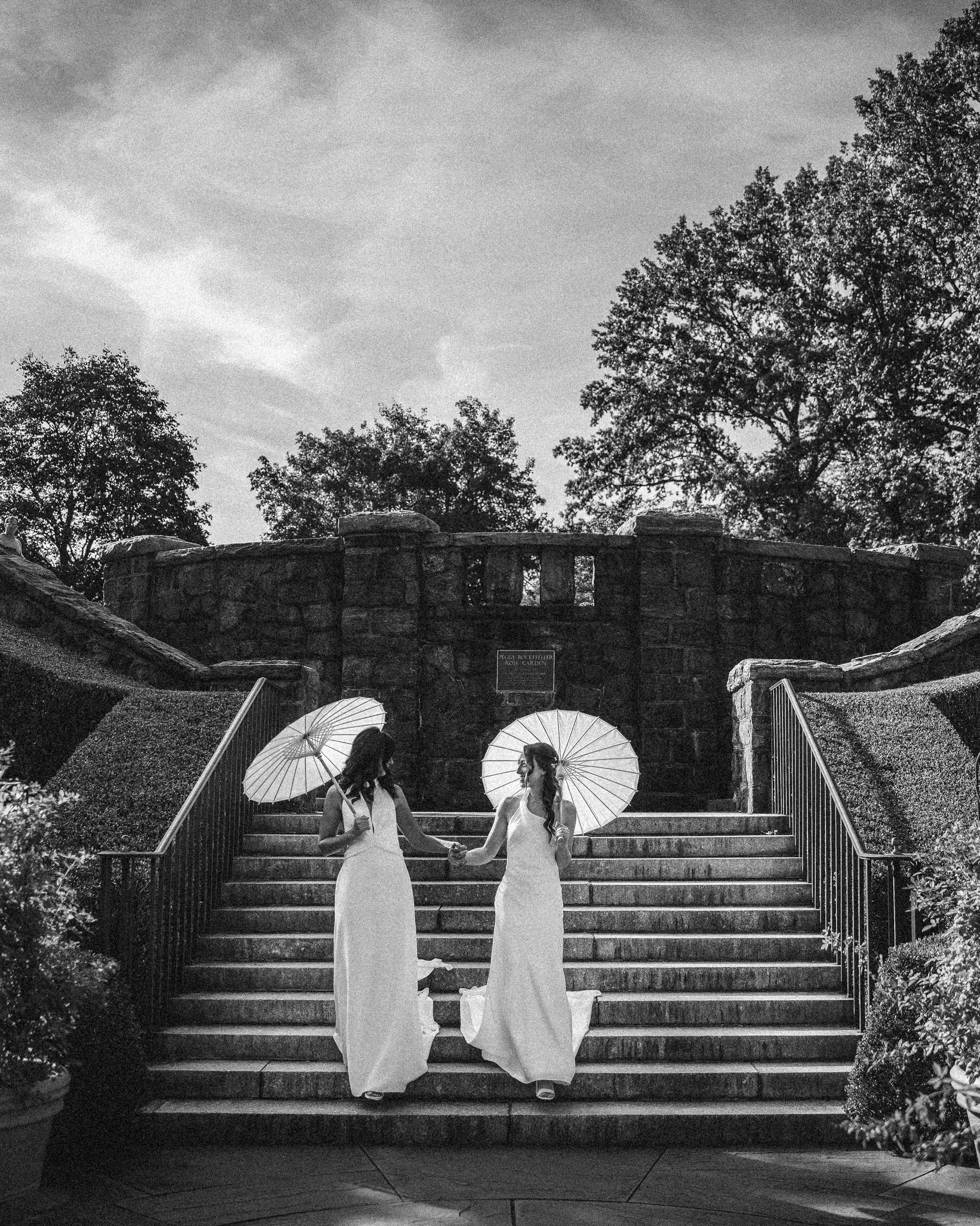 Two brides in white dresses holding parasols ascend outdoor stone steps surrounded by trees and shrubs, with a stone wall visible at the top—a timeless black and white photo evoking a New York Botanical Garden wedding.