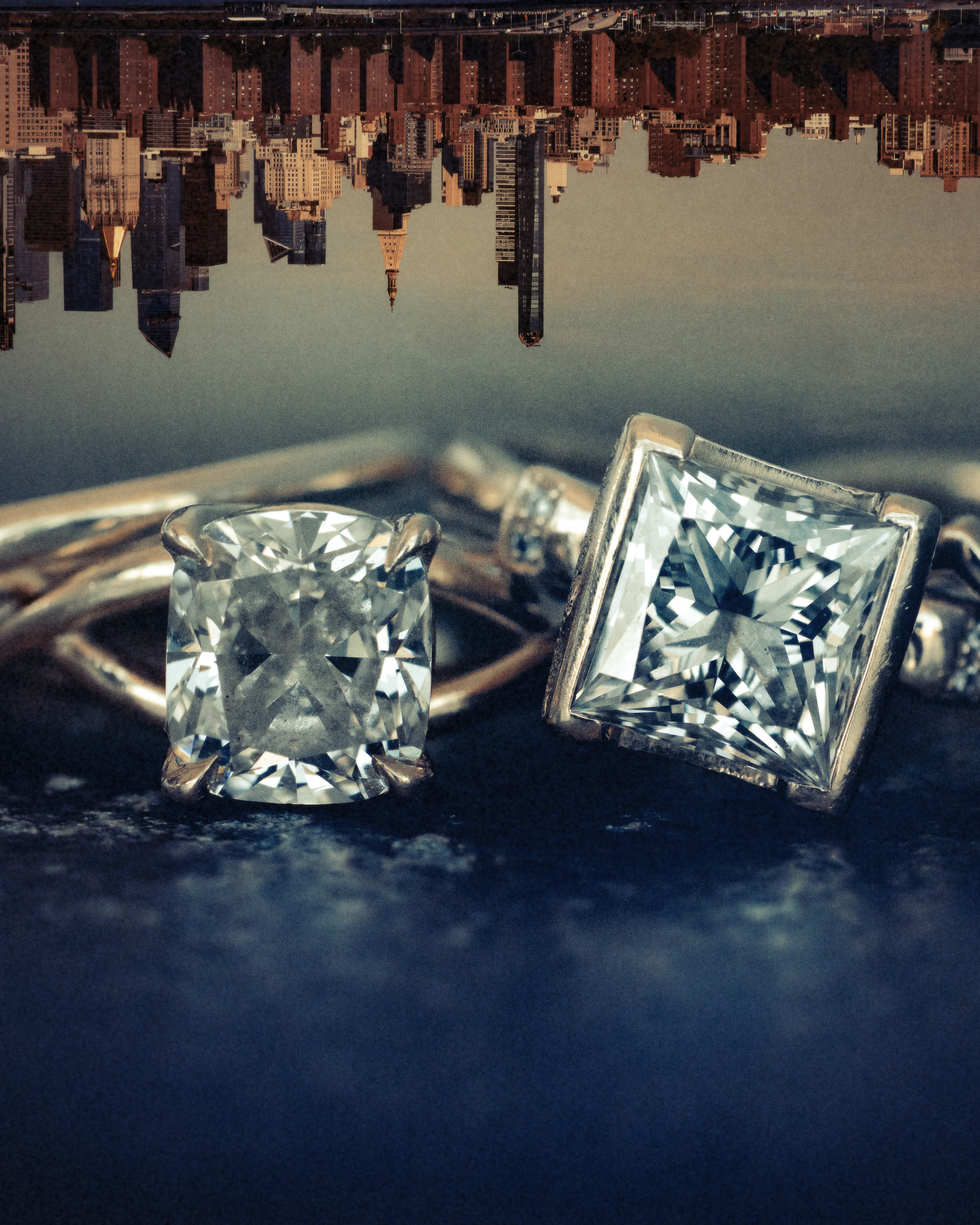 Two square-cut diamond rings on a dark surface with an upside-down city skyline in the background, evoking the elegance of a New York Botanical Garden wedding.