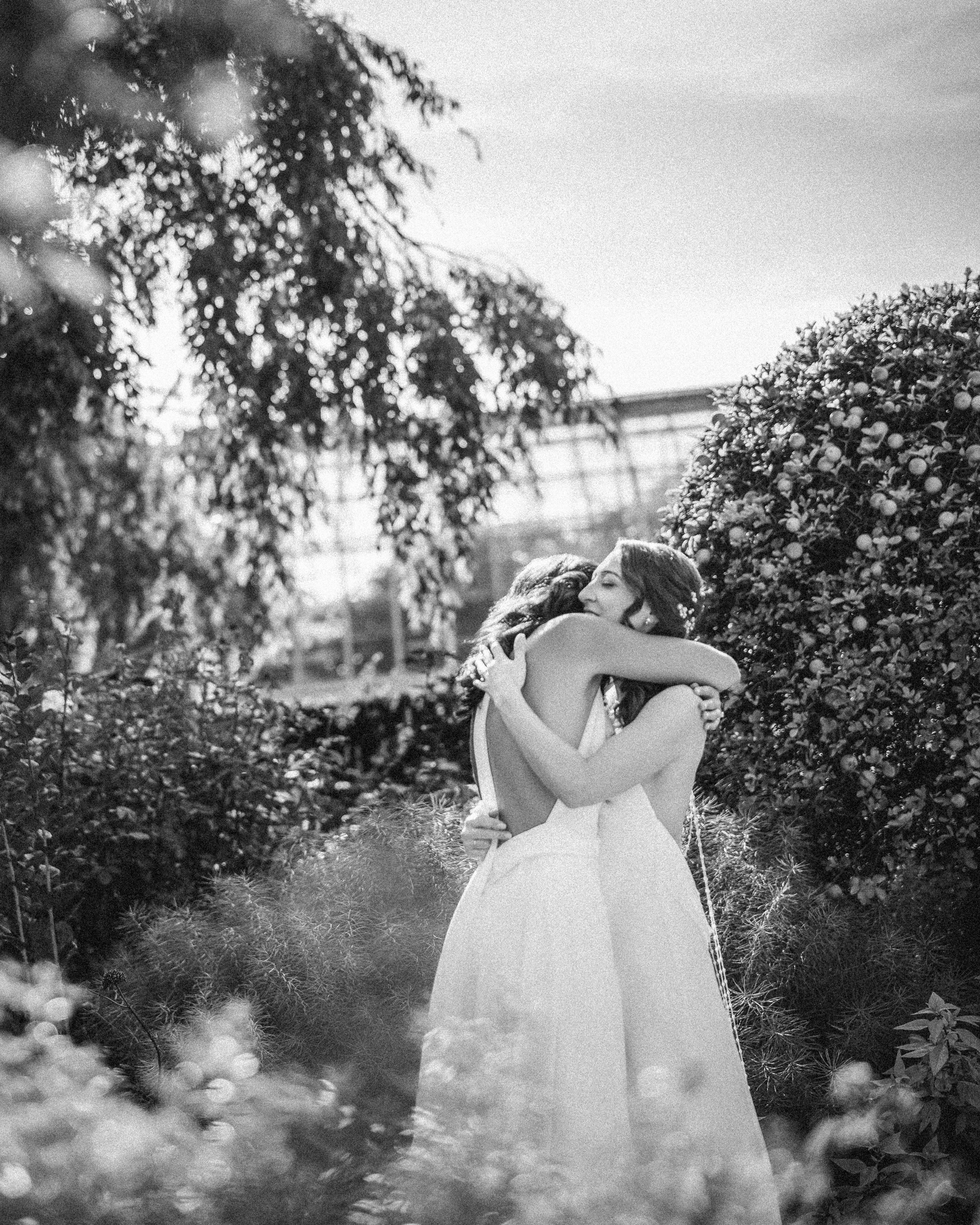 Two brides in white dresses embrace outdoors amid foliage and sunlight at the New York Botanical Garden wedding, captured in a timeless black-and-white photograph.