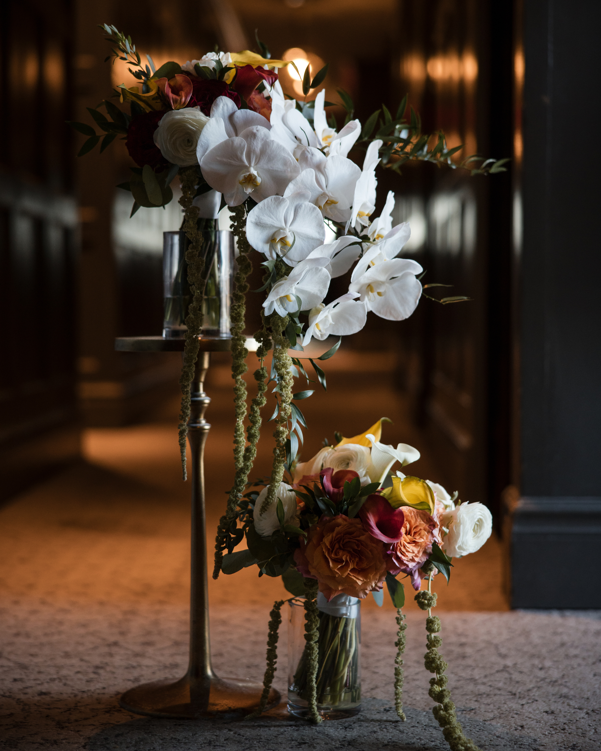 Two floral arrangements with white orchids, orange roses, and green foliage displayed on a brass stand and in a glass vase capture the elegance of a New York Botanical Garden wedding, set in a dimly lit hallway.
