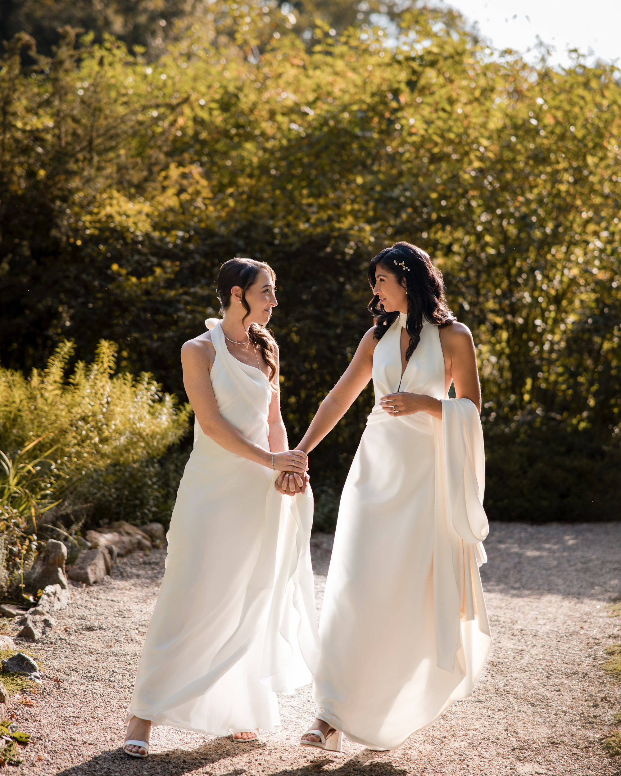Two brides in white dresses stand outdoors on a gravel path at their New York Botanical Garden wedding, holding hands and smiling at each other, surrounded by lush green foliage and sunlight.