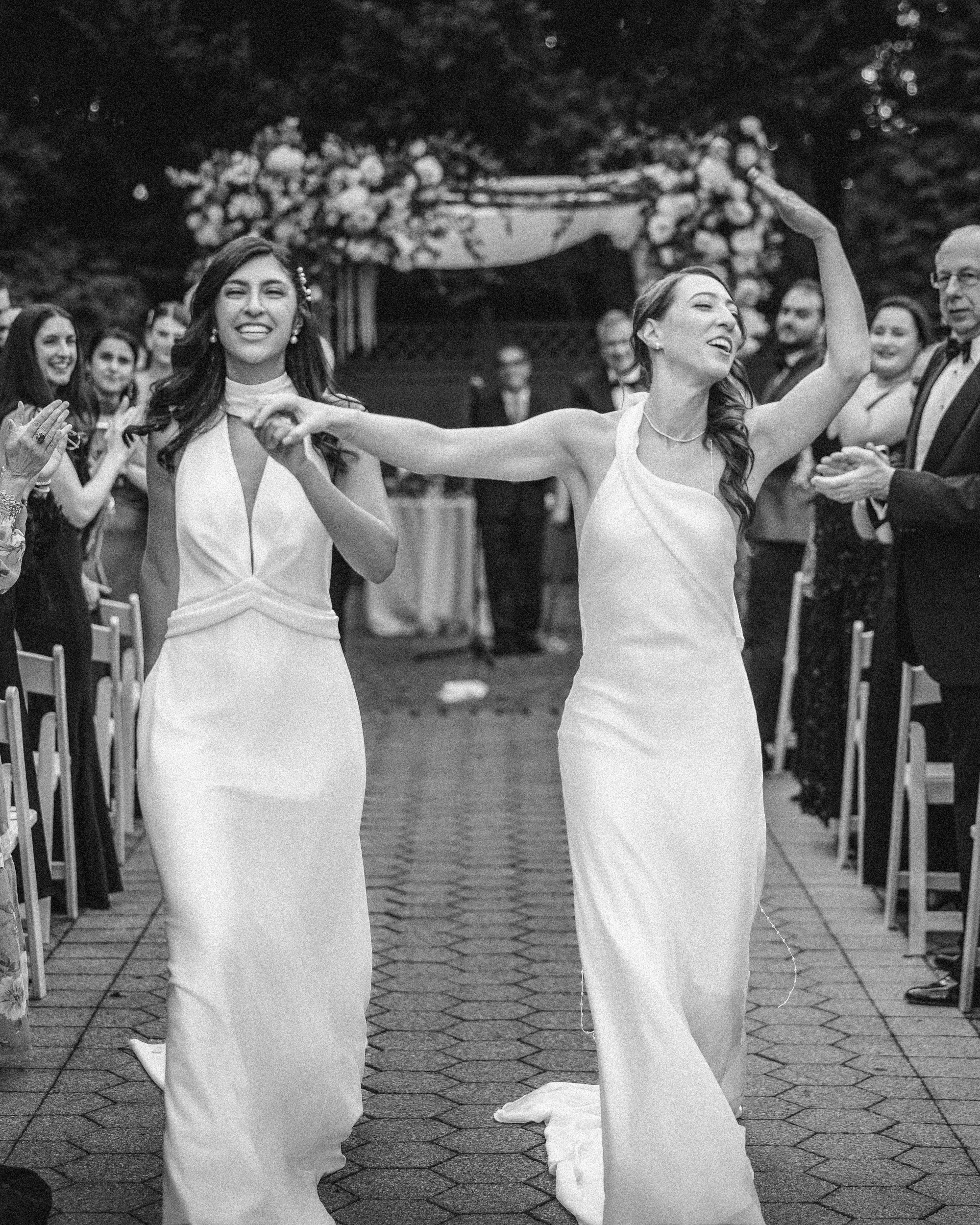 Two brides in white dresses walk down an outdoor aisle at their New York Botanical Garden wedding, smiling and holding hands, surrounded by applauding guests.