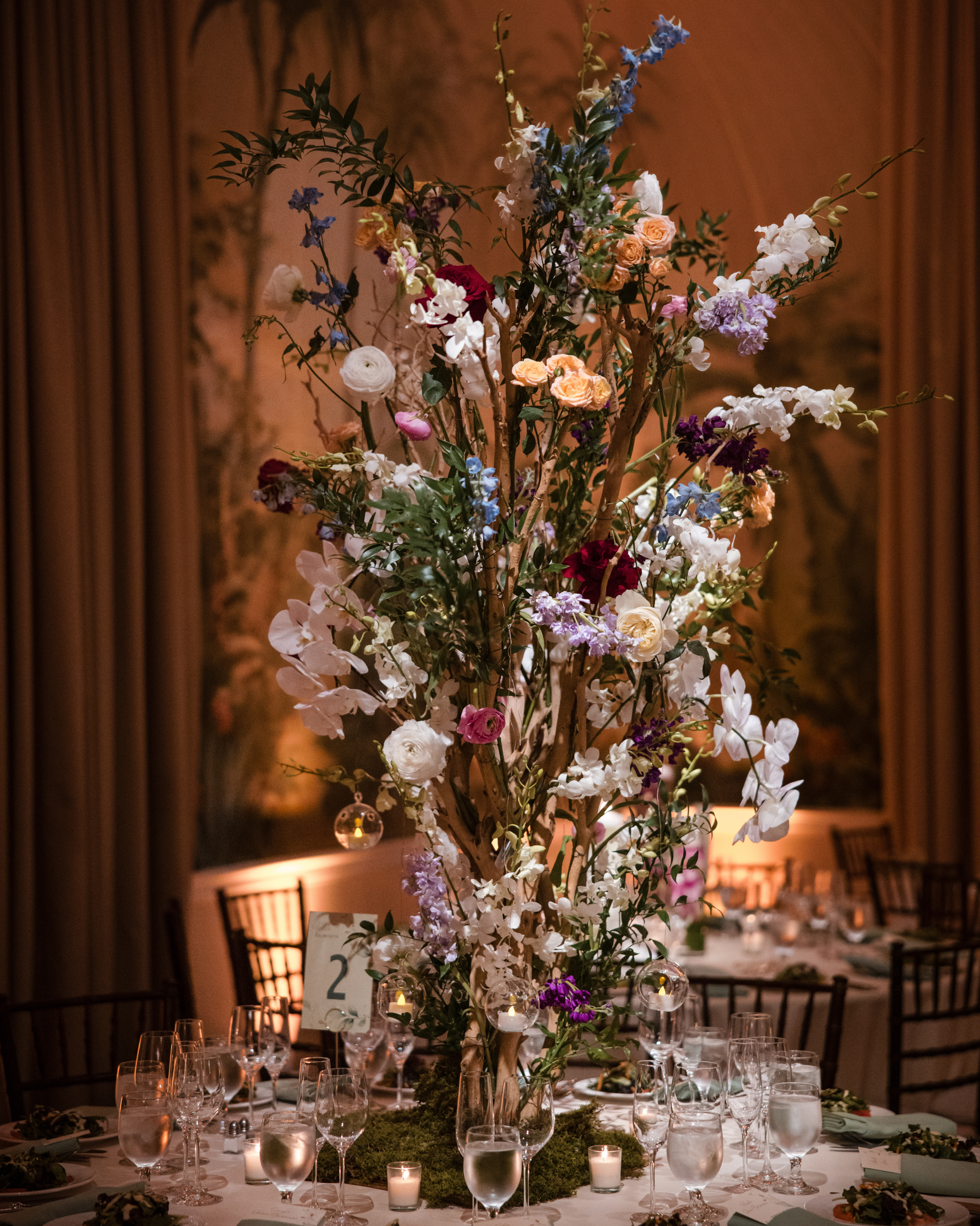 Tall floral centerpiece with multicolored flowers and branches adorns a round dining table set with glassware, plates, and candles in a dimly lit, elegant room—perfect for a New York Botanical Garden wedding.