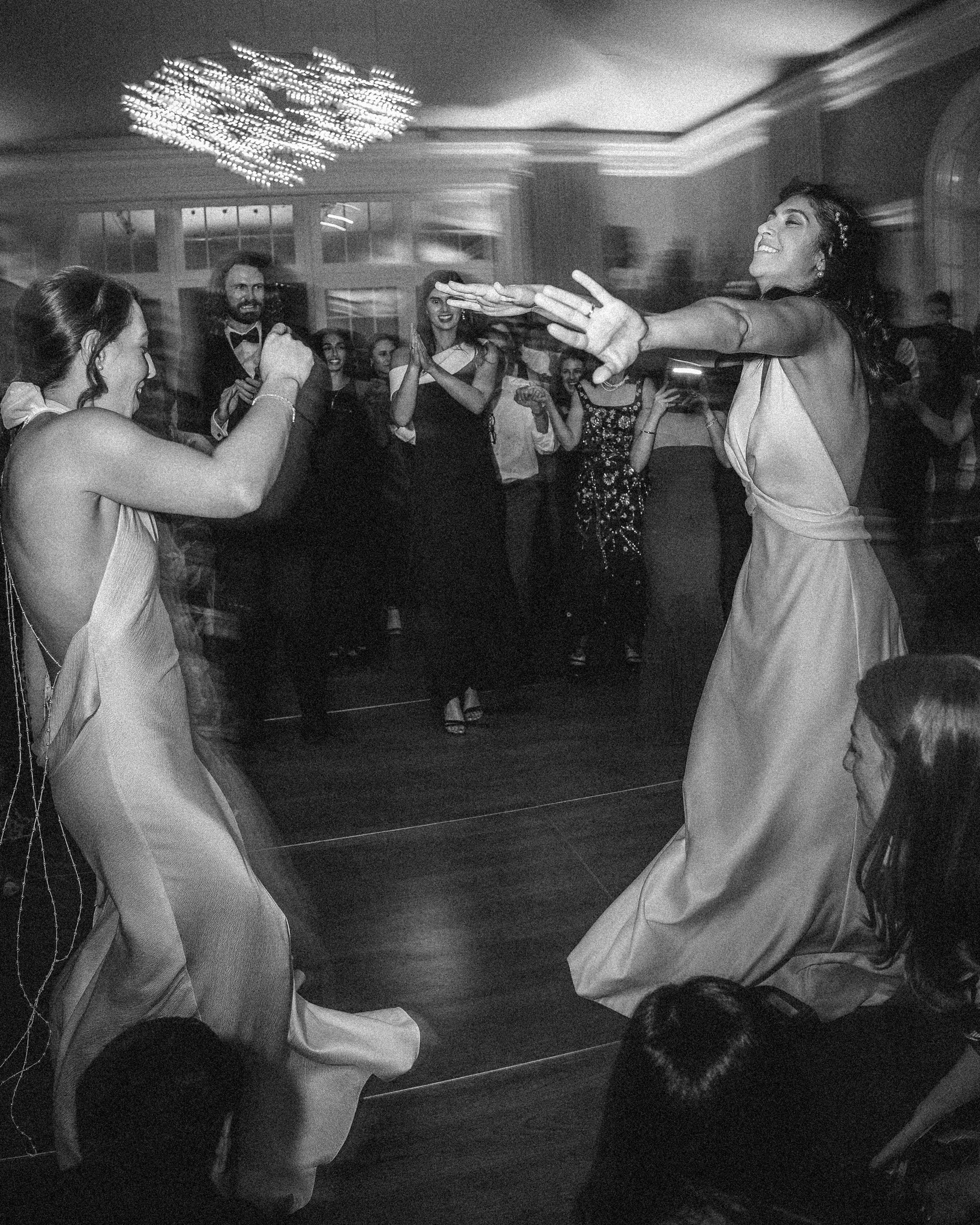 Two brides in formal dresses dance energetically together on a crowded dance floor at an indoor New York Botanical Garden wedding, surrounded by onlookers watching and smiling.