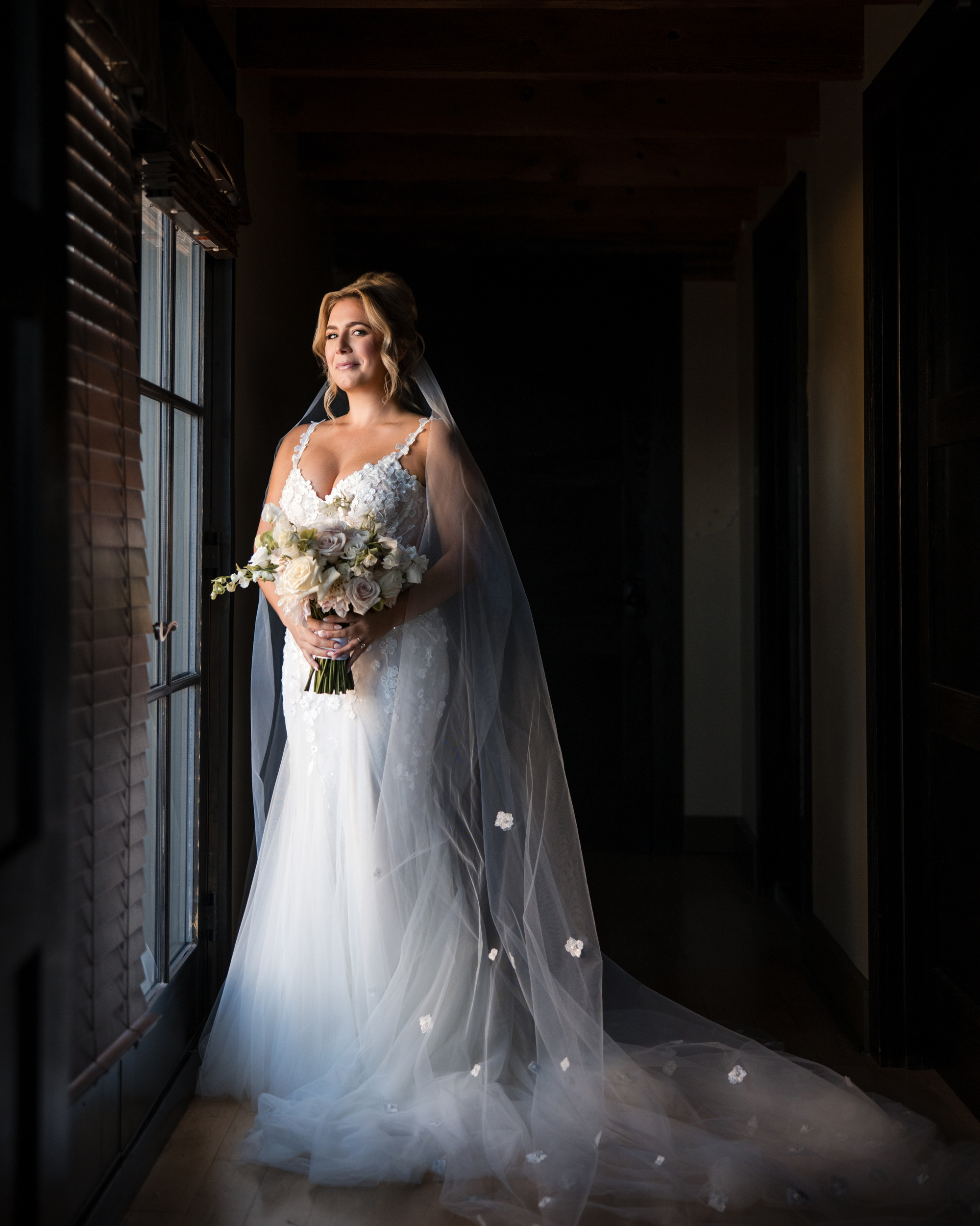 A bride in a white wedding dress and veil stands indoors by a window at the Lake House Inn, holding a bouquet of flowers and looking outside.