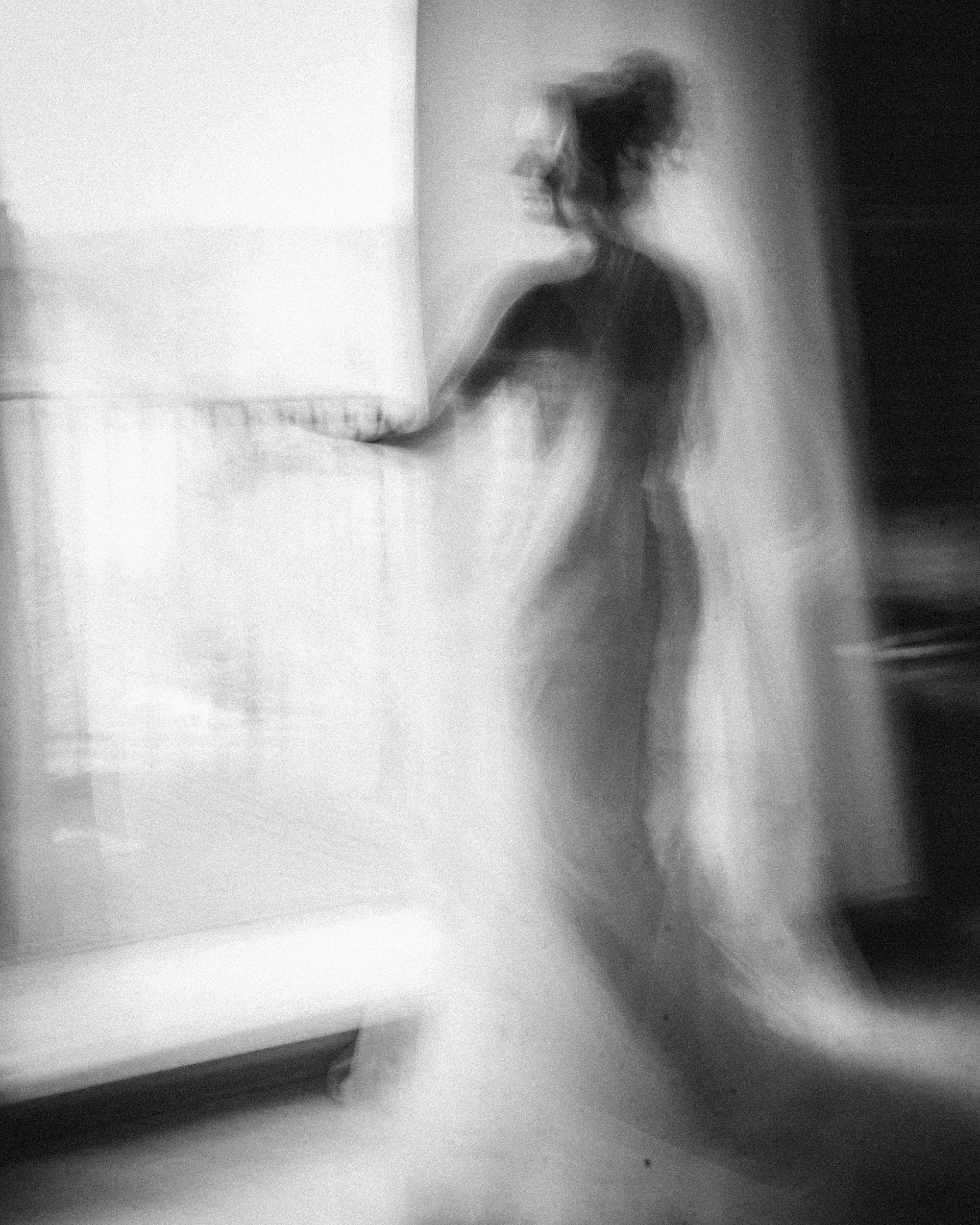 A blurred, black-and-white photo of a person in a gown standing indoors near a window at the Lake House Inn wedding, with light streaming in from outside.