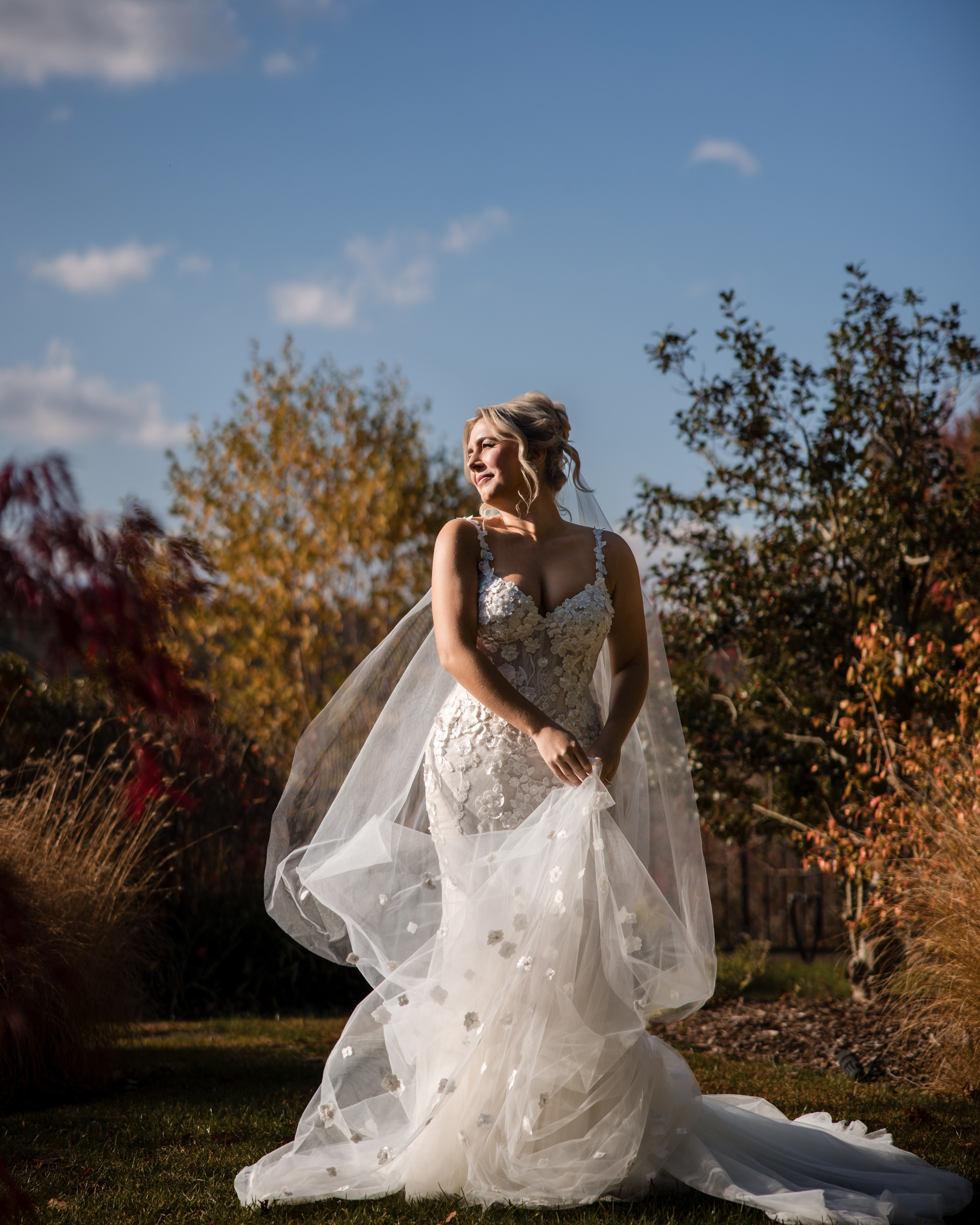 A bride in a white, fitted wedding dress with a sheer veil stands outdoors on grass at her Lake House Inn wedding, surrounded by autumn trees under a blue sky.