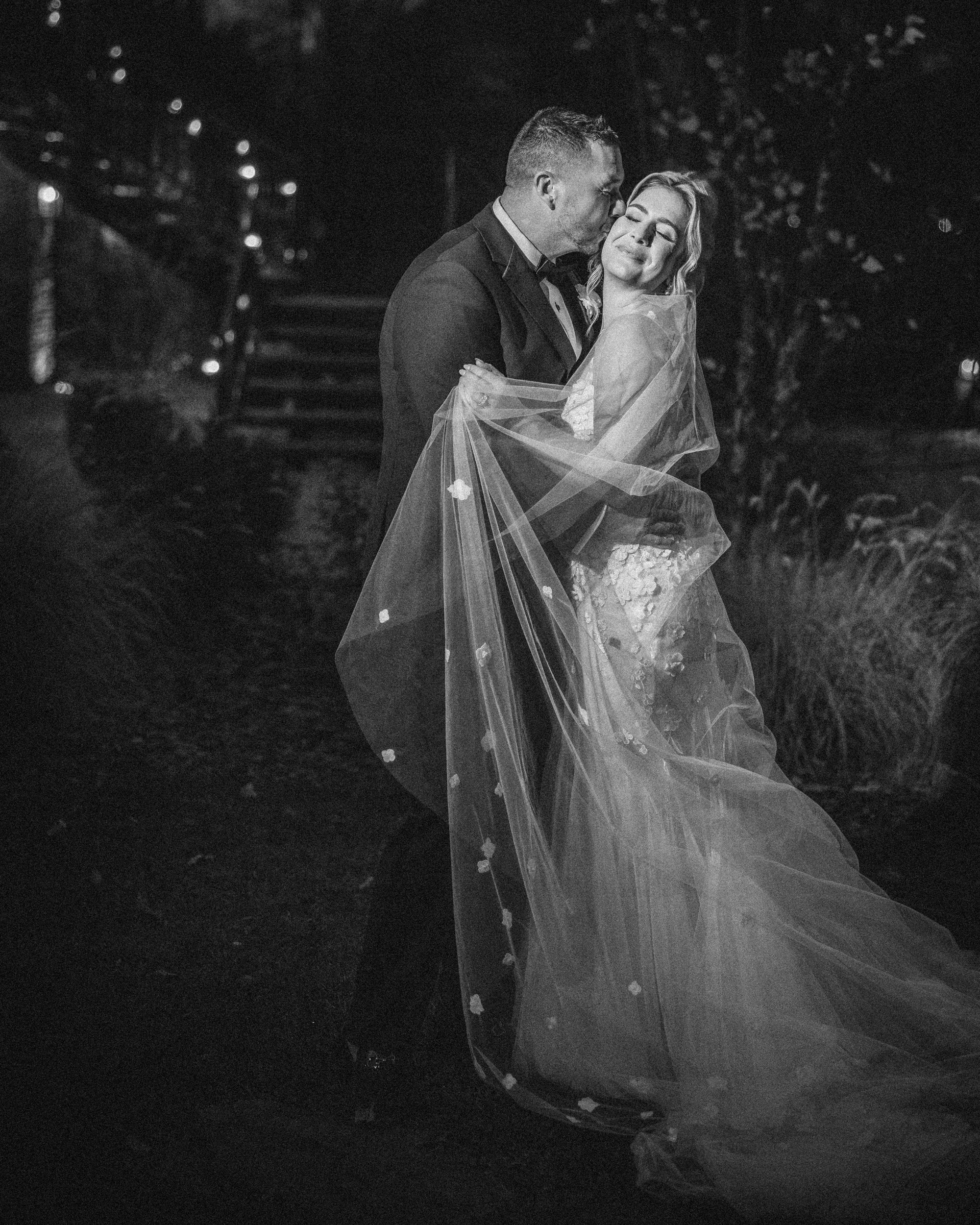 A bride and groom stand outdoors at night during their Lake House Inn wedding; the groom kisses the smiling bride, who holds her veil, both dressed in formal wedding attire.