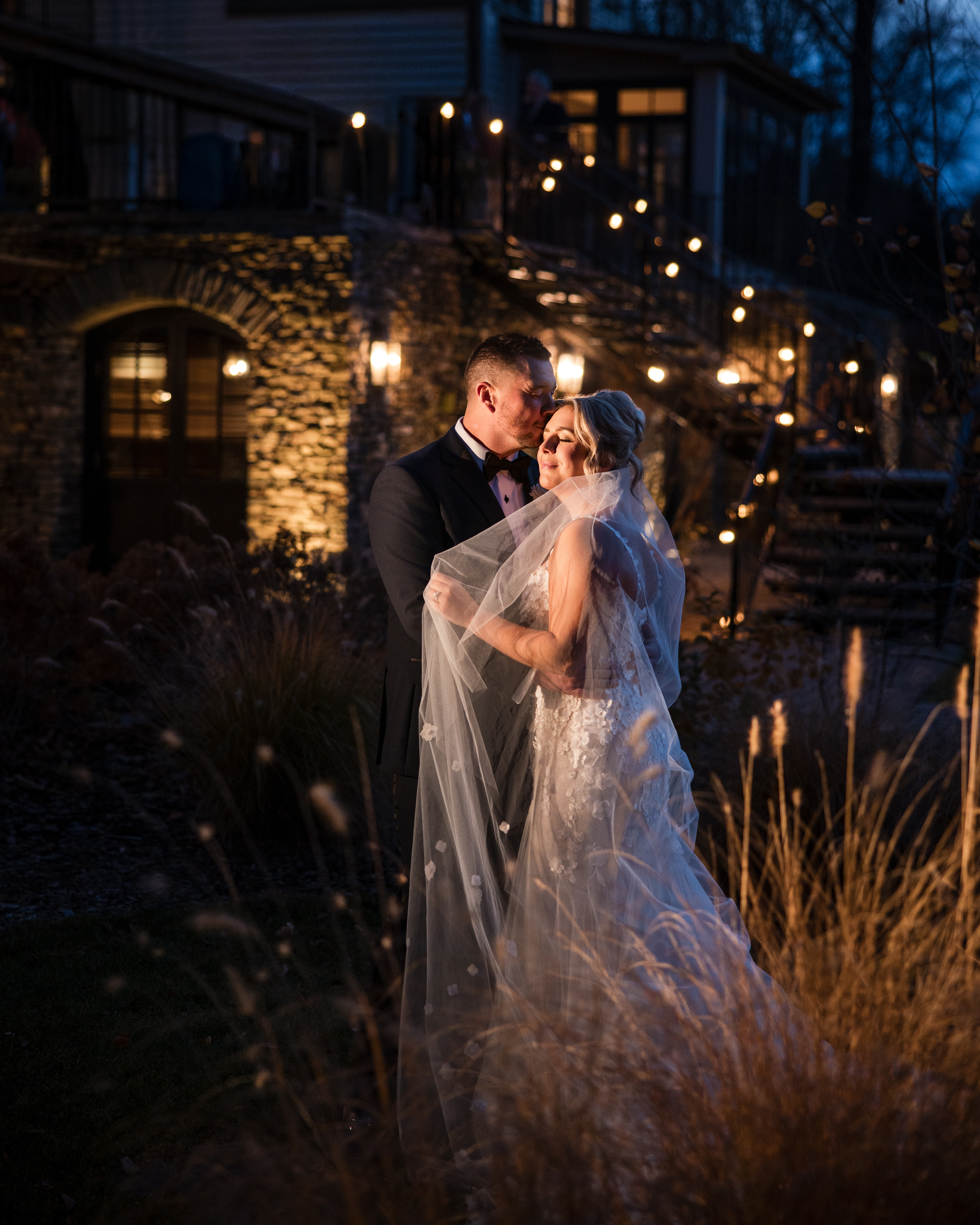 A bride and groom embrace outdoors at night, illuminated by warm lights, with a stone building and staircase in the background—a romantic Lake House Inn wedding moment.