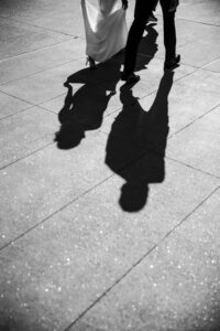 Two people stroll on a sunlit pavement near Castell Rooftop Lounge, their full shadows trailing behind. Only their legs and part of a dress are visible, hinting at an elegant St. Patrick's Cathedral wedding celebration.