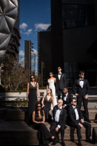 A group of eight people in formal black and white attire pose outdoors in bright sunlight at Castell Rooftop Lounge, with modern buildings in the background, celebrating after a St. Patrick’s Cathedral wedding.