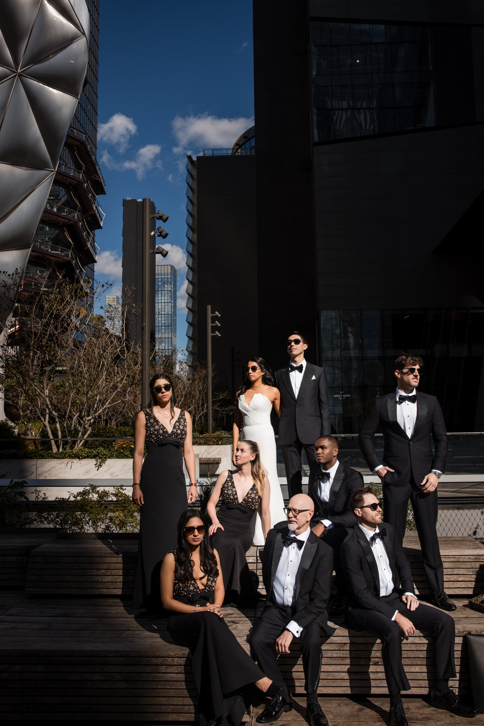A group of eight people in formal black and white attire pose outdoors in bright sunlight at Castell Rooftop Lounge, with modern buildings in the background, celebrating after a St. Patrick’s Cathedral wedding.