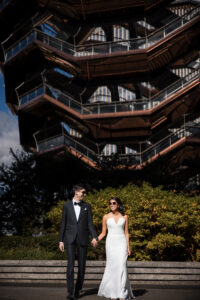 A couple in formal wedding attire holds hands and walks outdoors in front of the Vessel, after celebrating their st patricks cathedral wedding in New York City.