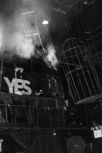 Black and white photo of a theater stage at a House of Yes wedding, featuring a large "YES" sign, stage equipment, an empty cage, visible EXIT sign, and smoke rising near the ceiling.