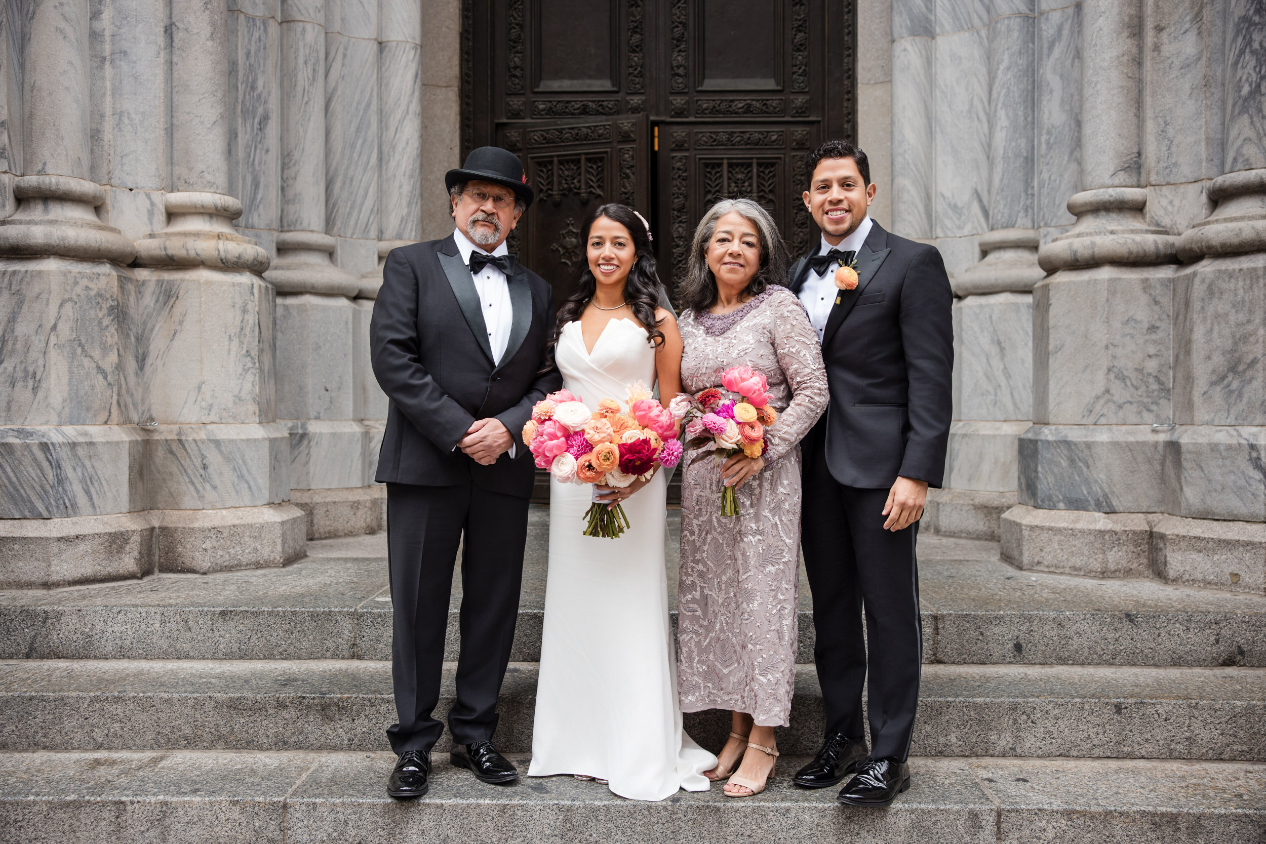 Four people dressed formally stand on stone steps near St. Patrick's Cathedral, with two holding bouquets. The ornate building’s large doors and marble columns create a stunning wedding backdrop, perfect for a Castell Rooftop Lounge celebration after.