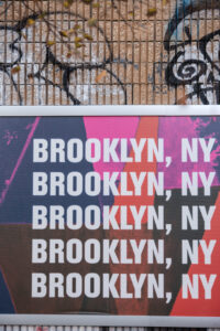A billboard displays the words "BROOKLYN, NY" repeated four times in bold white text against a colorful, abstract background, evoking the vibrant spirit you'd find at a House of Yes wedding, with graffiti on the wall above.
