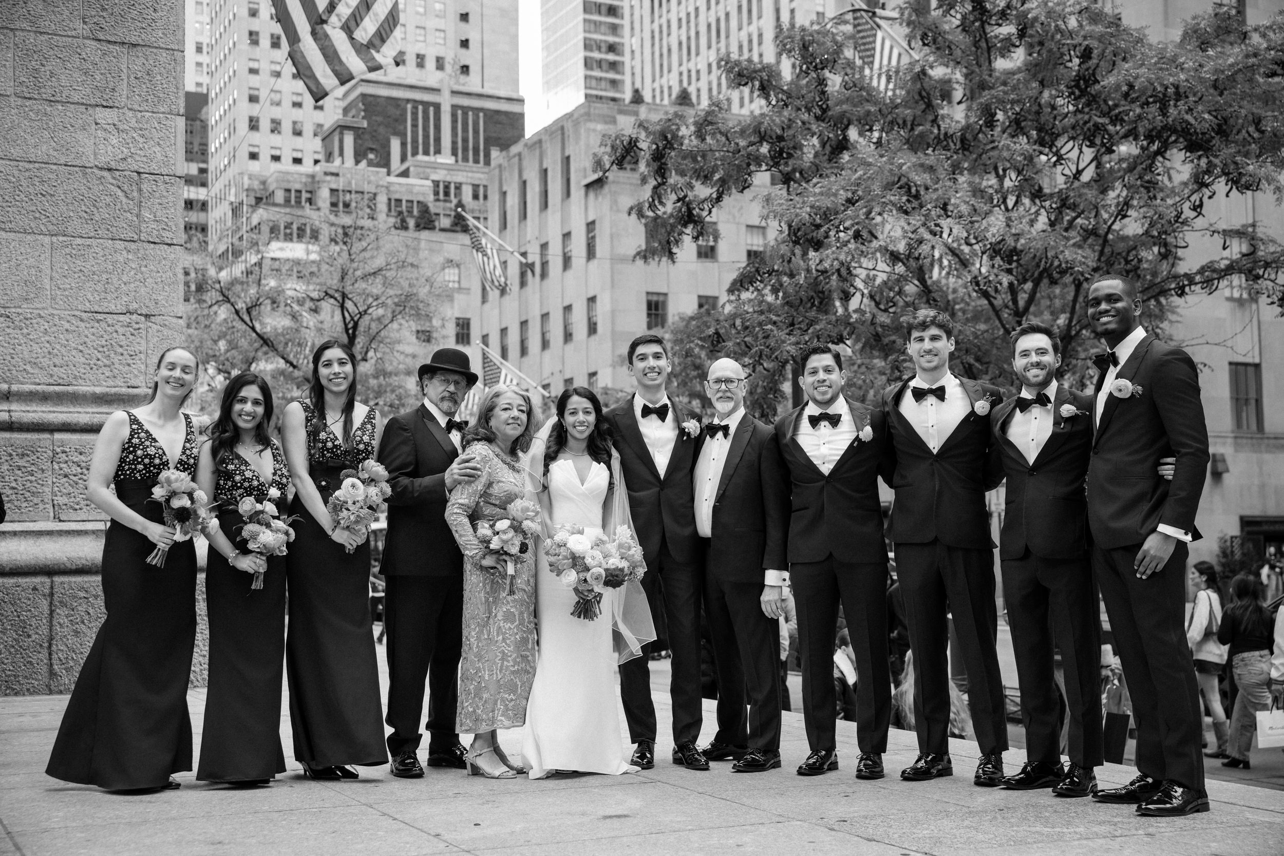 A wedding party of twelve, including the bride and groom, pose together outdoors in formal attire with the city skyline behind them near Castell Rooftop Lounge after their St. Patrick's Cathedral wedding.