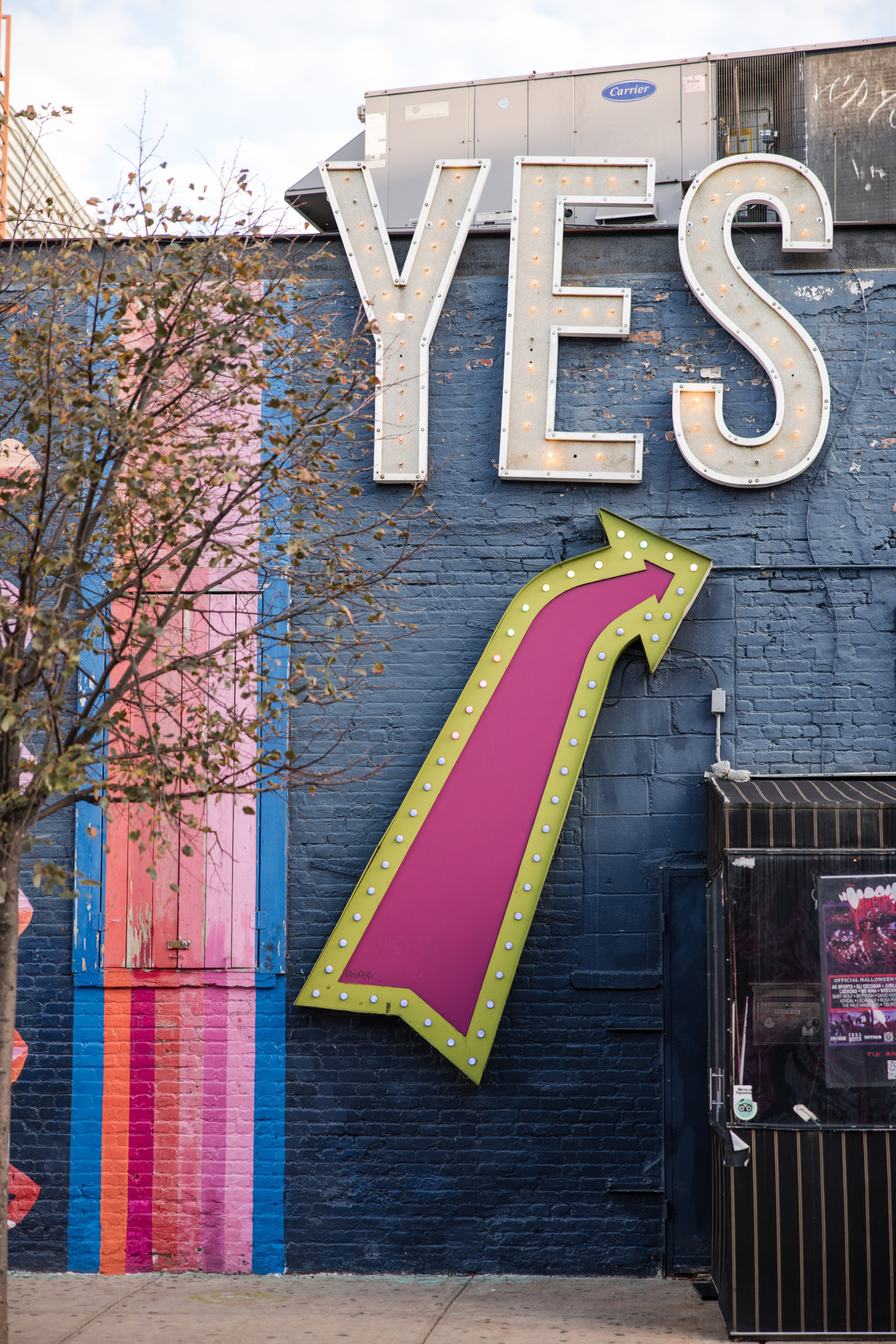 Large marquee letters spelling "YES" are mounted on a blue brick wall above a pink and yellow arrow sign—perfect for capturing that House of Yes wedding magic—with a small tree and storefront in the foreground.