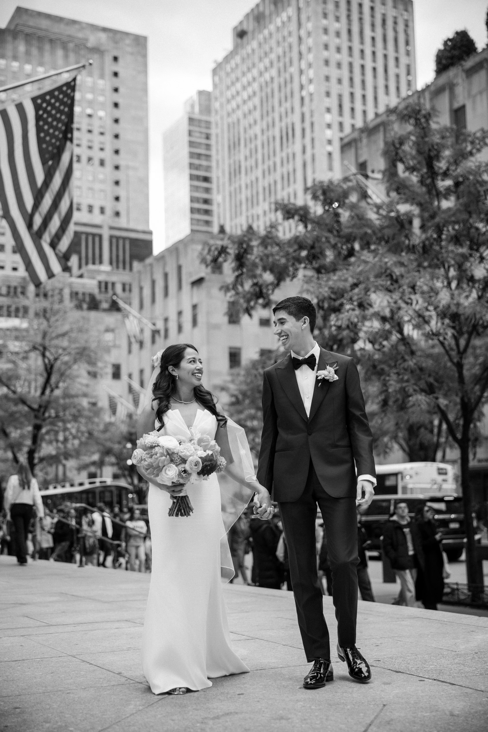 A bride and groom walk hand-in-hand on a city street in formal wedding attire, with tall buildings, the American flag, and the iconic Castell Rooftop Lounge visible in the background.