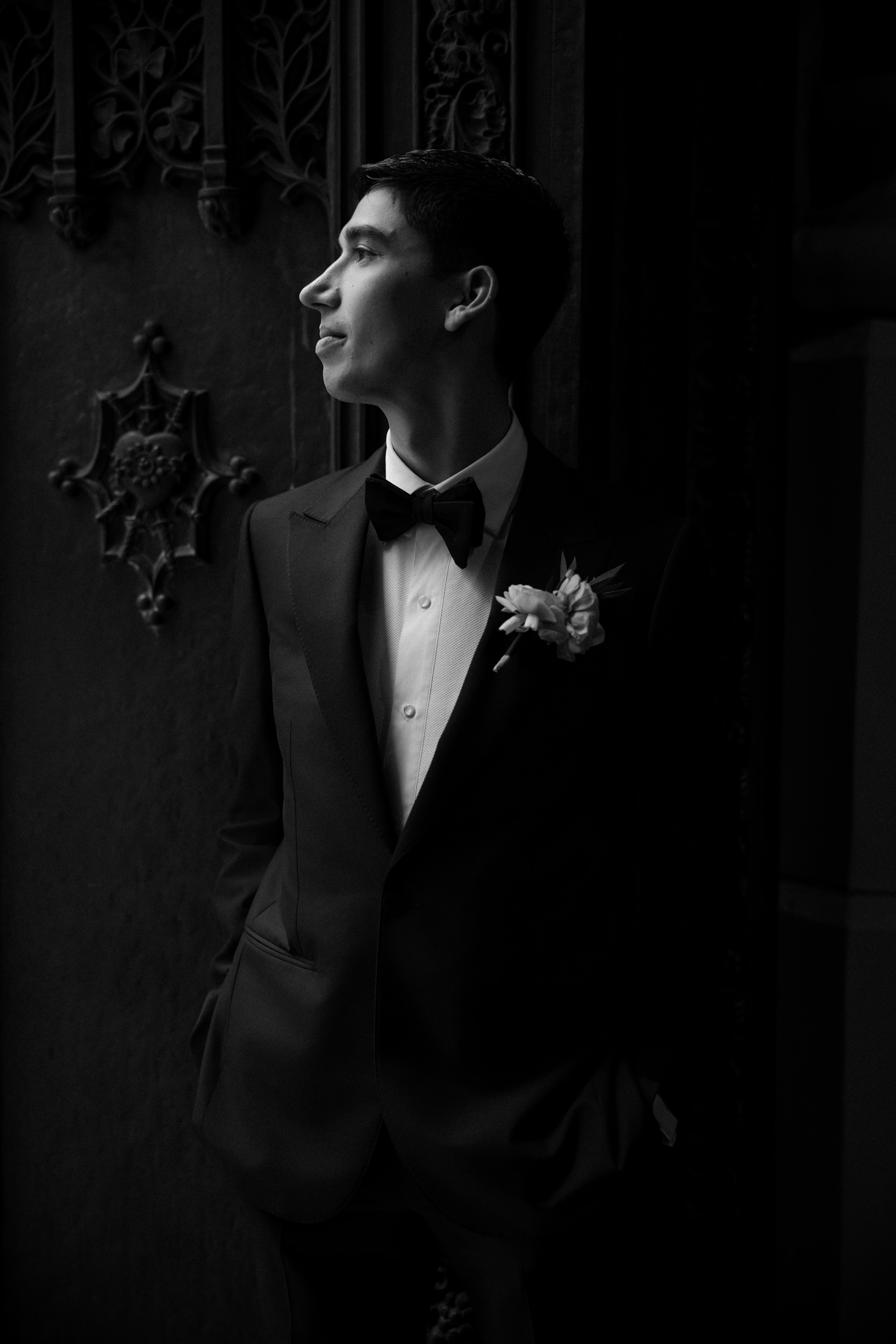 A young man in a tuxedo with a boutonnière stands near an ornate door at the Castell Rooftop Lounge, looking to the side in low light, recalling moments from a stunning St. Patrick's Cathedral wedding.