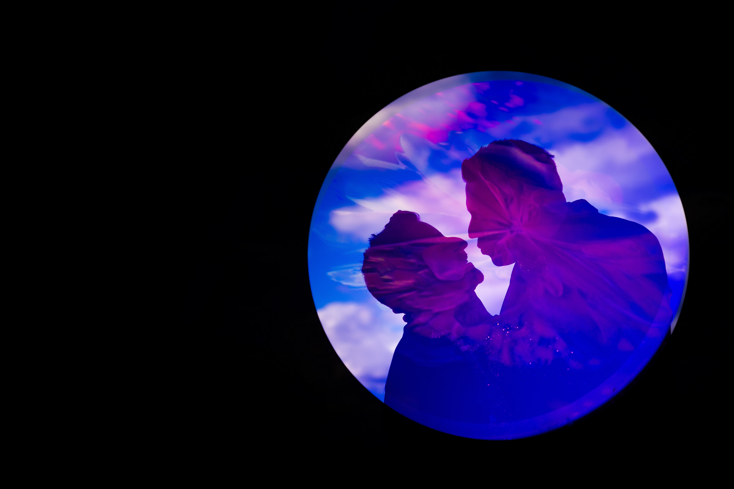 Silhouettes of two people facing each other inside a circular frame with a purple and blue abstract background, set against a black backdrop—perfect for capturing the unique vibe of a House of Yes wedding.
