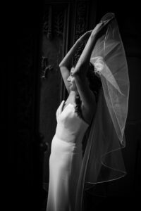 A woman in a wedding dress stands indoors at Castell Rooftop Lounge, lifting her veil above her head as light from the side illuminates her, evoking the elegance of a St. Patrick's Cathedral wedding.