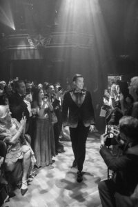 A man in a tuxedo walks confidently down a lit aisle as an audience on either side watches and takes photos, capturing the vibrant atmosphere of a House of Yes wedding in a formal, indoor setting.