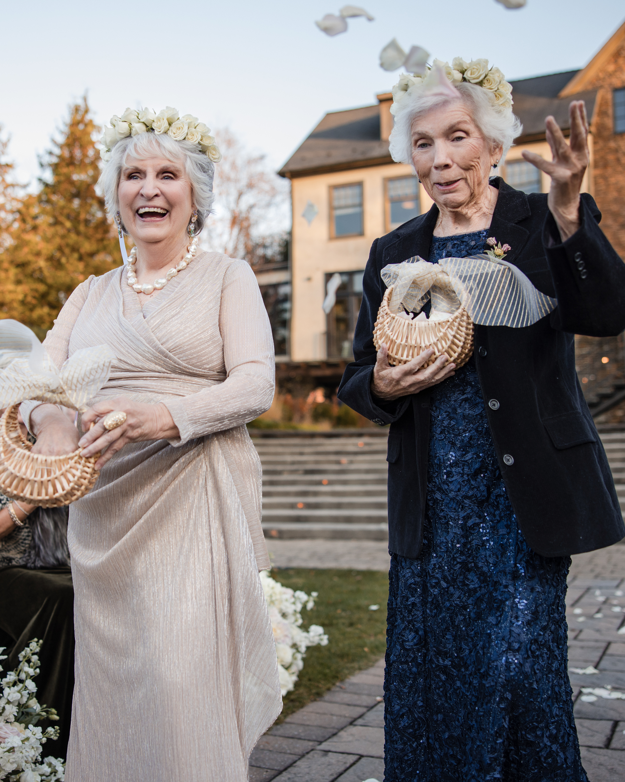Two grandmas at a Lake House Inn wedding, dressed in formal attire and floral crowns, hold baskets and smile outdoors as white doves fly above them near the steps.