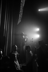 A singer performs on stage with a microphone in hand at a House of Yes wedding, facing an audience; several people stand nearby in the dimly lit, dramatic scene.