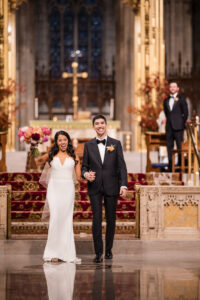 A bride and groom, both smiling, stand hand in hand at the altar after their St. Patrick’s Cathedral wedding. The bride holds a bouquet of flowers while a groomsman looks on—moments before celebrating at the Castell Rooftop Lounge.