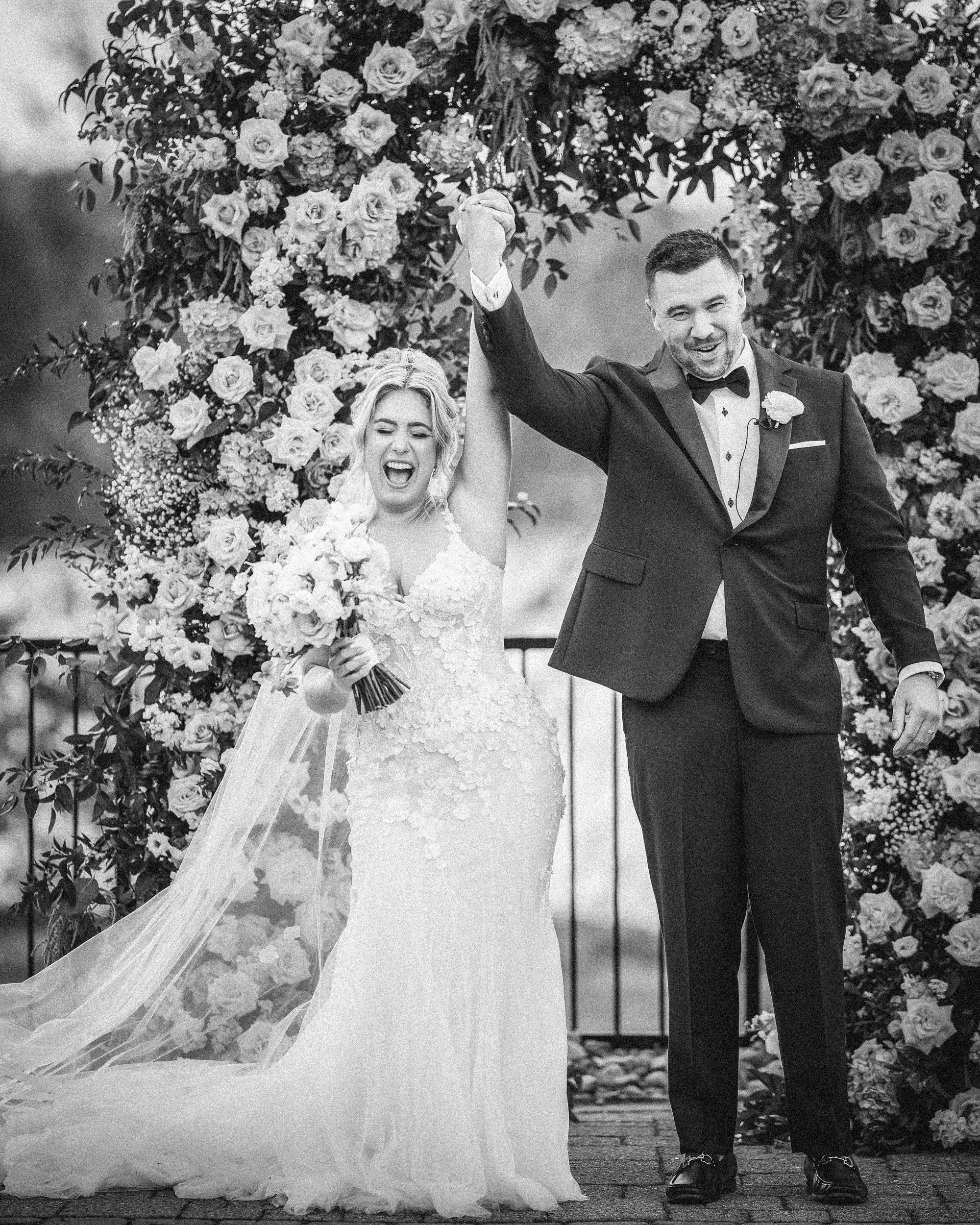 A bride and groom stand under a floral arch at their Lake House Inn wedding, holding hands and raising them in celebration on their special day.