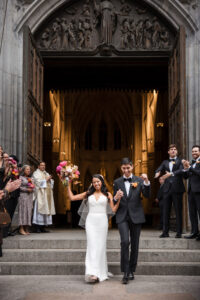 A bride and groom walk down the steps of St. Patrick's Cathedral, smiling and holding hands, as guests celebrate and clap—moments before their chic reception at Castell Rooftop Lounge.