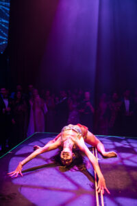 A woman in a performance costume bends backward on stage under purple lighting at a House of Yes wedding, with an audience watching in the background.