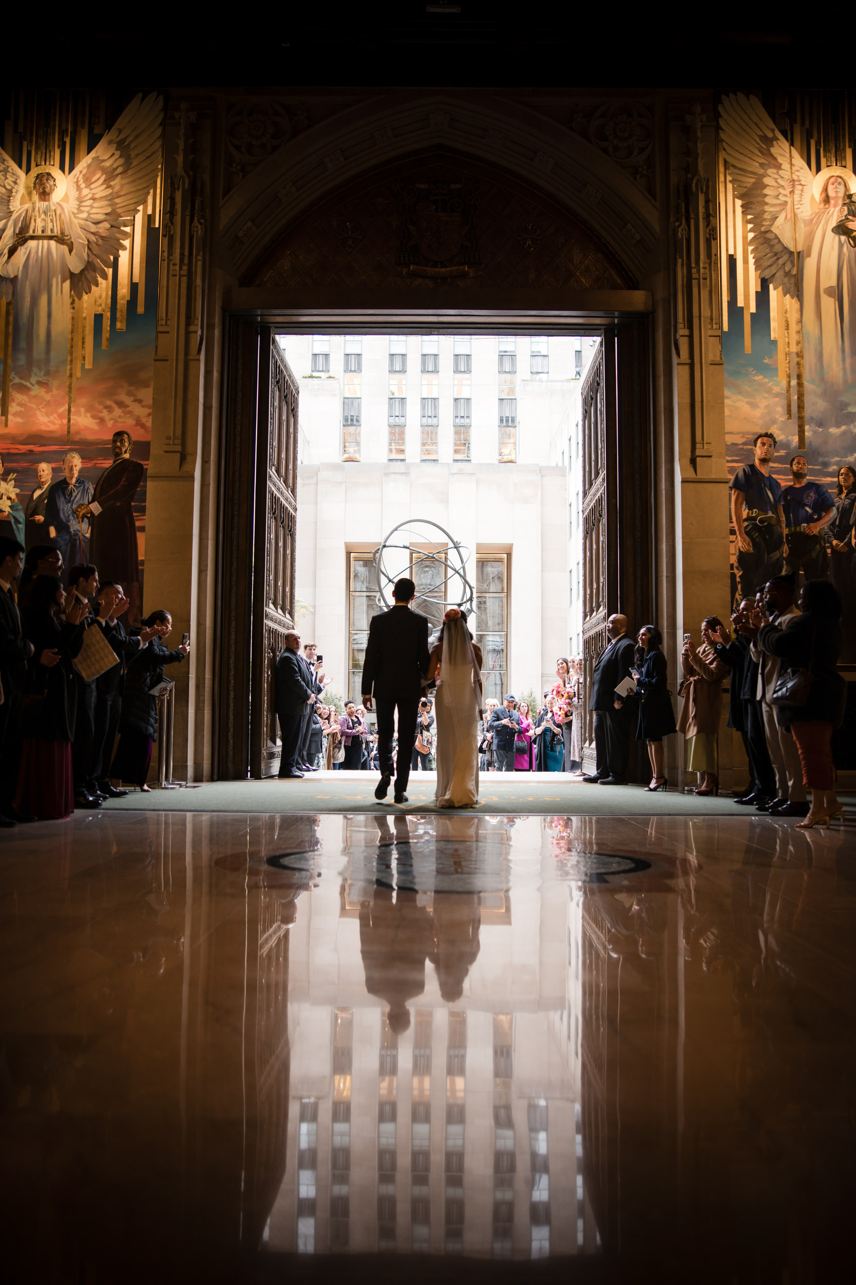 A bride and groom stand hand in hand at the entrance of a grand church, reminiscent of a St. Patrick's Cathedral wedding, facing guests on either side and large murals of angels above the doors.