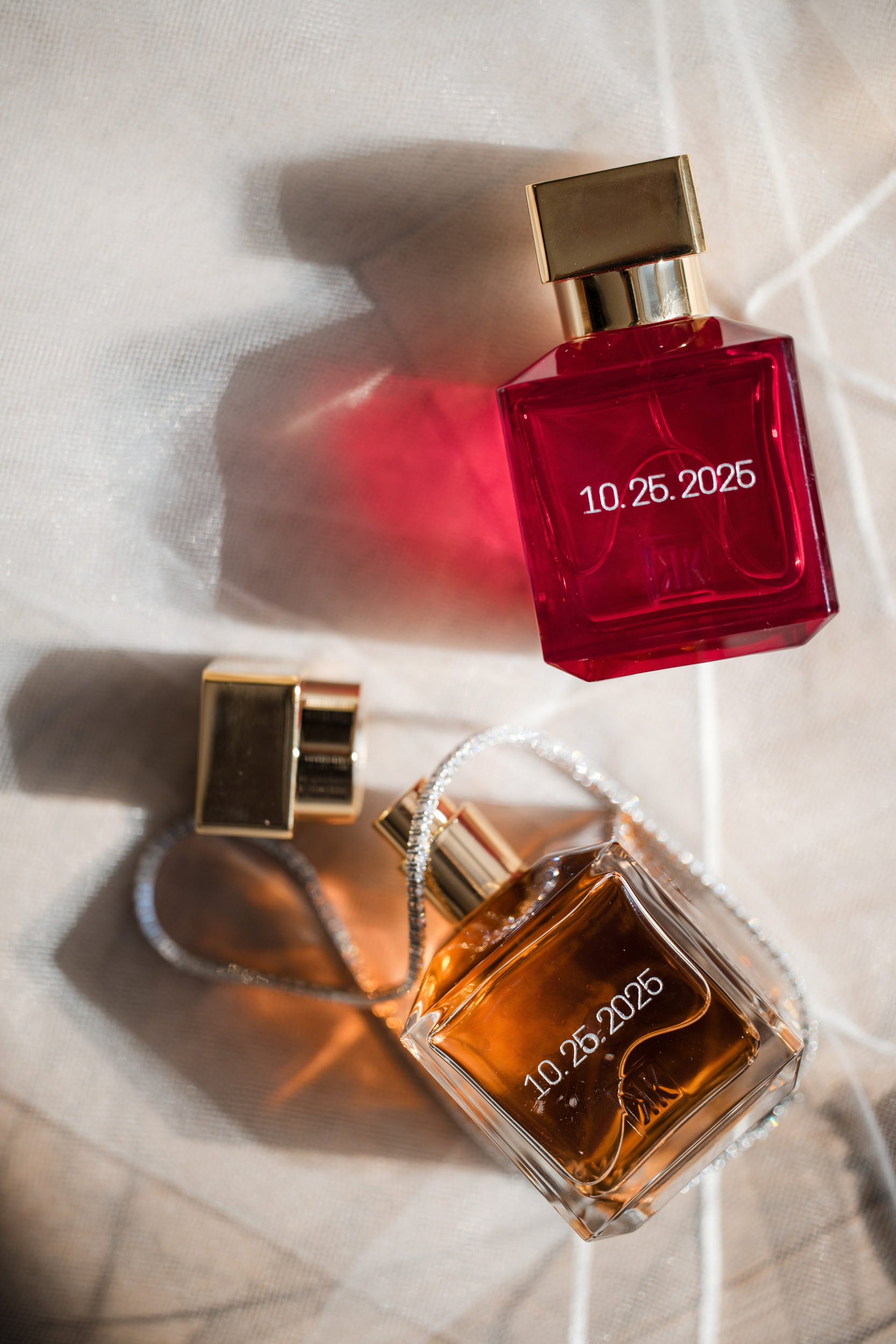 Two perfume bottles with gold caps, one red and one amber, both labeled "10.26.2026," rest on a light fabric surface with soft shadows—an elegant detail reminiscent of a Castell Rooftop Lounge St Patricks Cathedral wedding celebration.