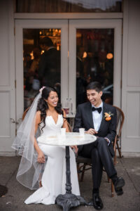 A bride and groom sit at a small round table outside a café, smiling and laughing together after their St. Patrick's Cathedral wedding. The bride wears a white dress and veil; the groom wears a black suit with a bow tie.
