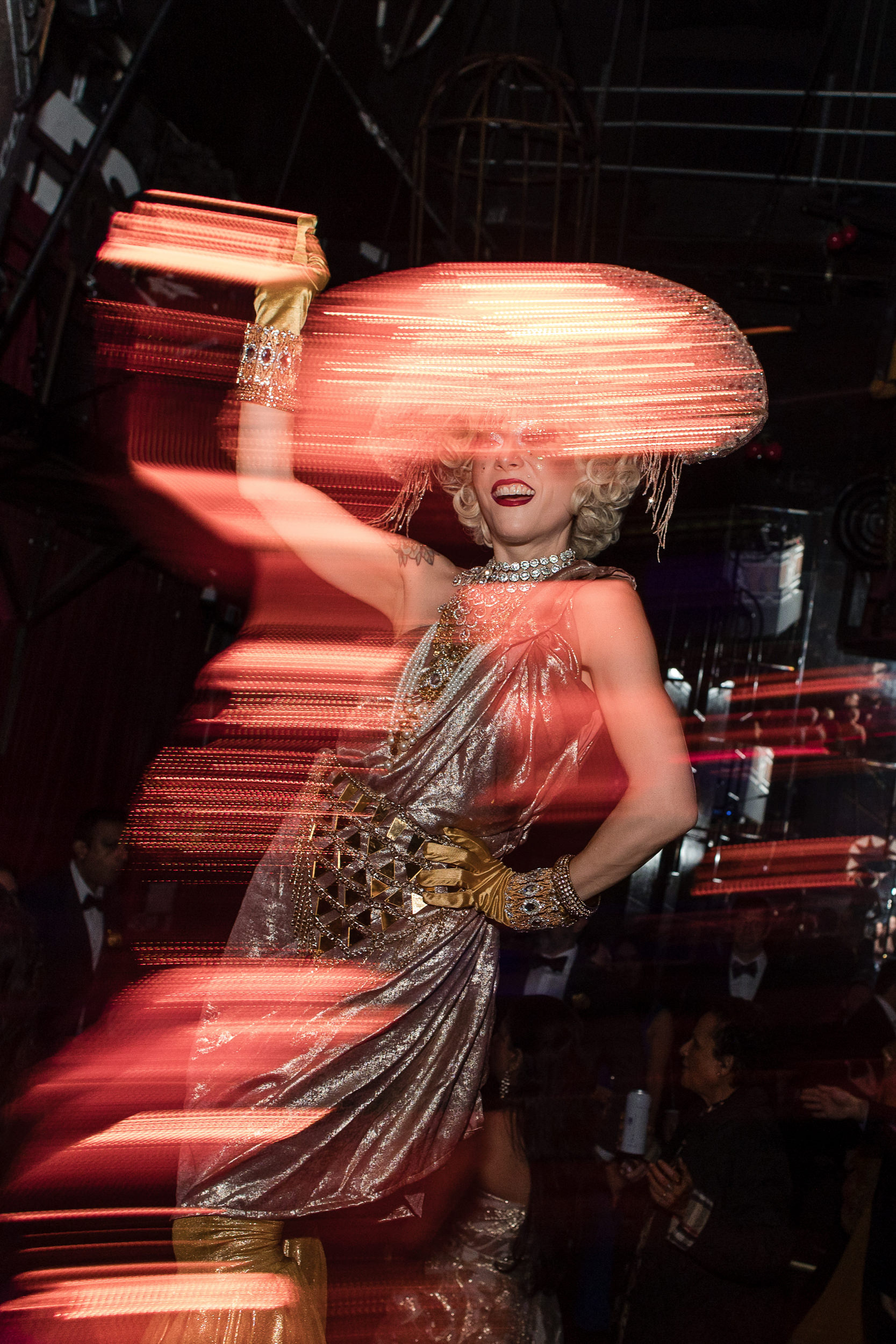 A person in a shiny, silver outfit and dramatic hat poses with a raised arm, surrounded by bright, streaking lights at a lively House of Yes wedding in a dark, indoor setting.