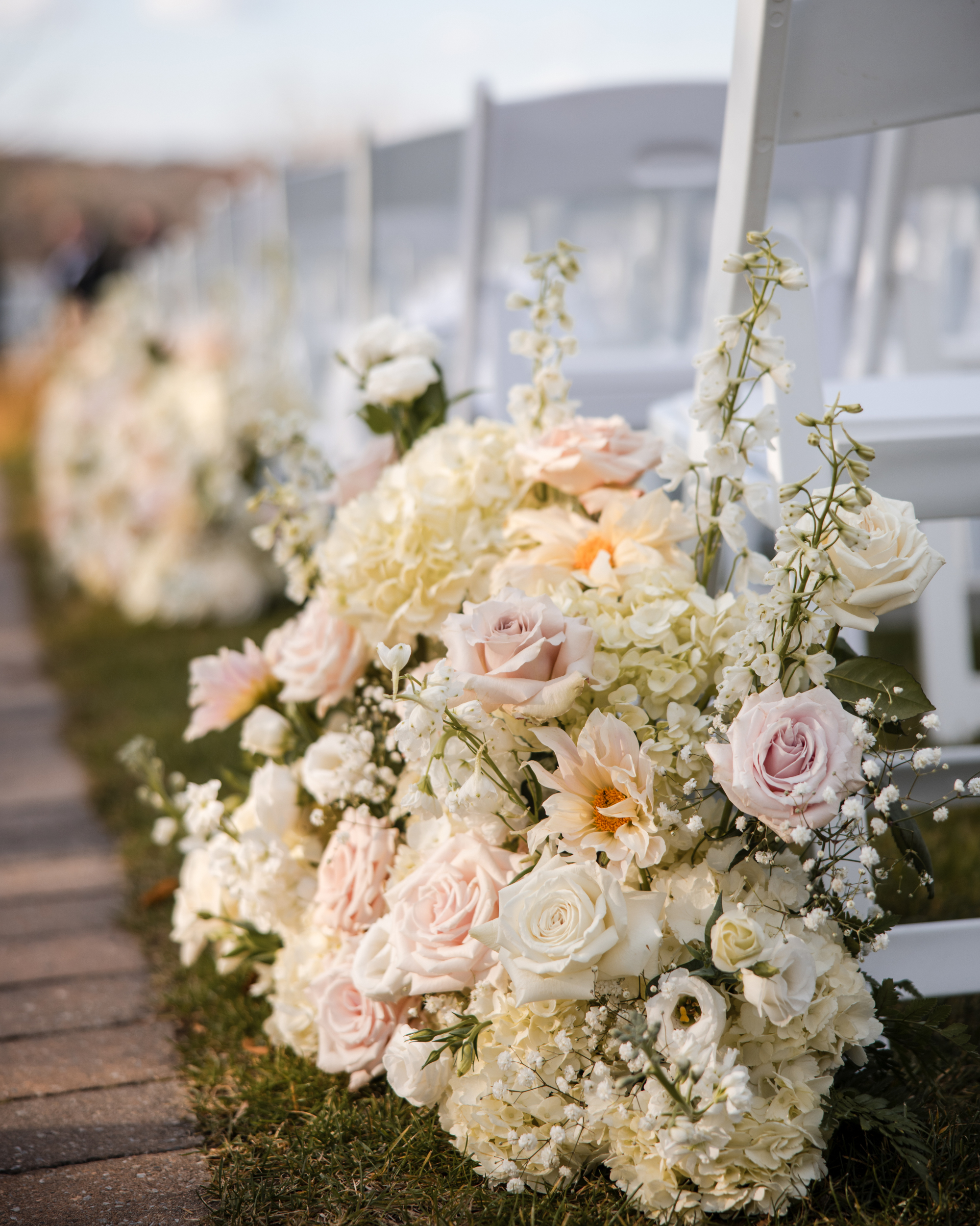 Rows of white folding chairs with arrangements of pale pink and white flowers line an outdoor aisle on grass, creating a romantic setting for a Lake House Inn wedding.