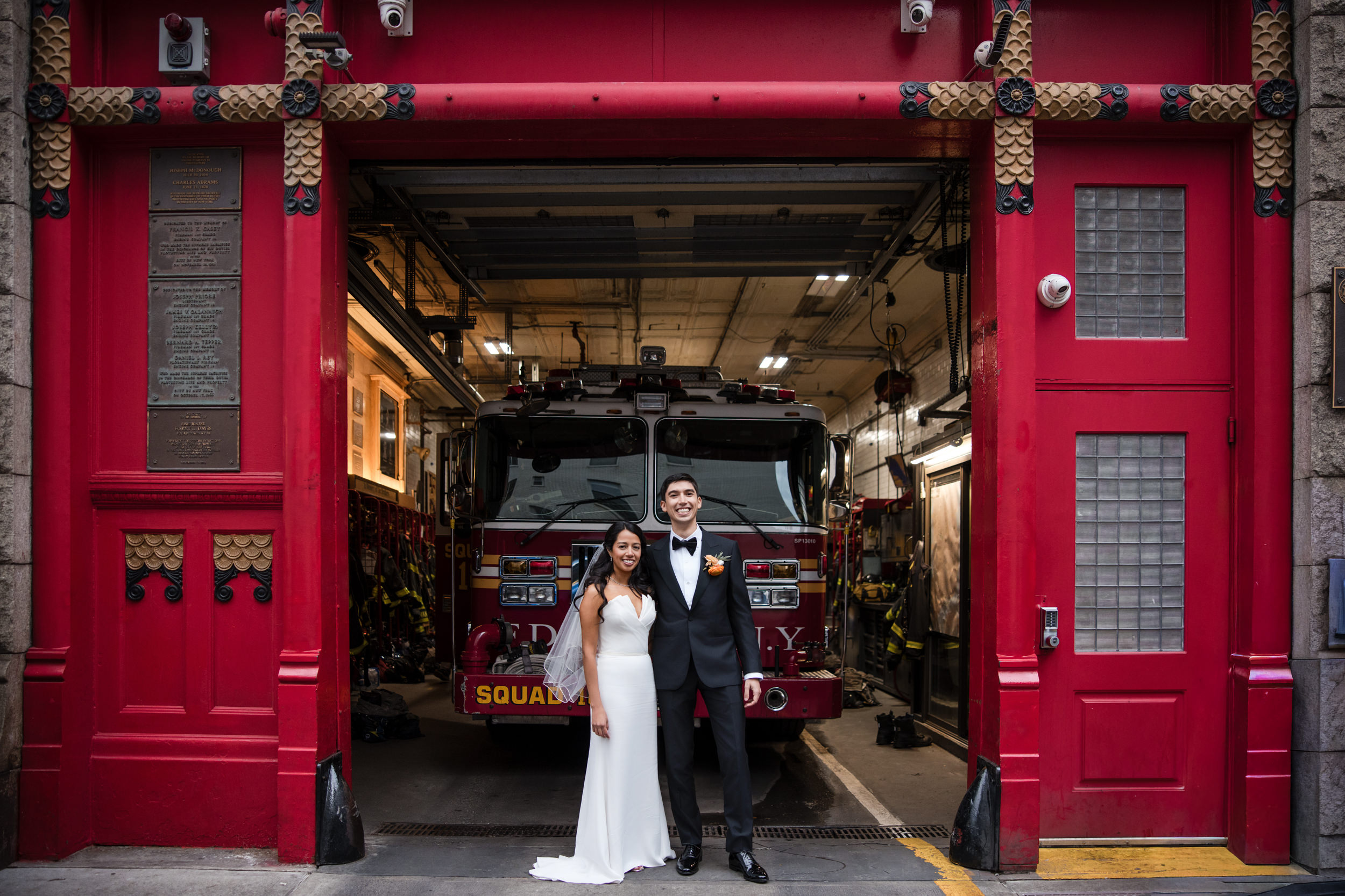 A bride and groom stand in front of a fire truck inside a fire station with large red doors, following their St. Patrick’s Cathedral wedding.