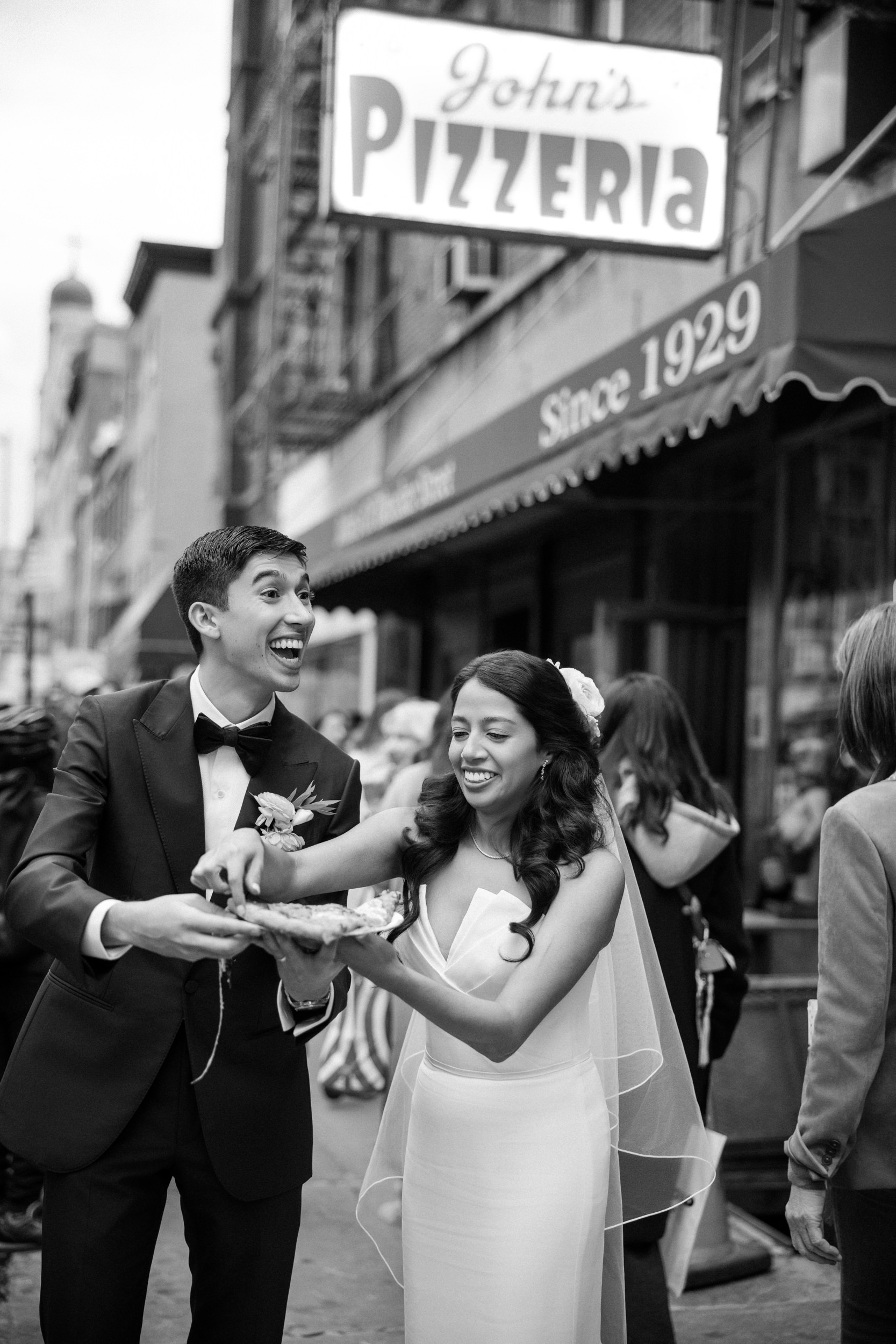 A bride and groom in formal attire laugh while sharing pizza in front of John's Pizzeria after their St. Patrick's Cathedral wedding, with other people in the background.