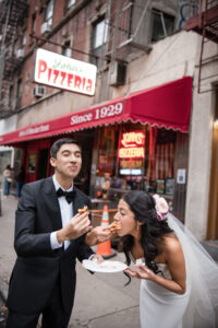 A bride and groom in formal attire eat pizza together outside a pizzeria with a red awning and neon sign, celebrating their St. Patrick’s Cathedral wedding on a lively city sidewalk before heading to Castell Rooftop Lounge.