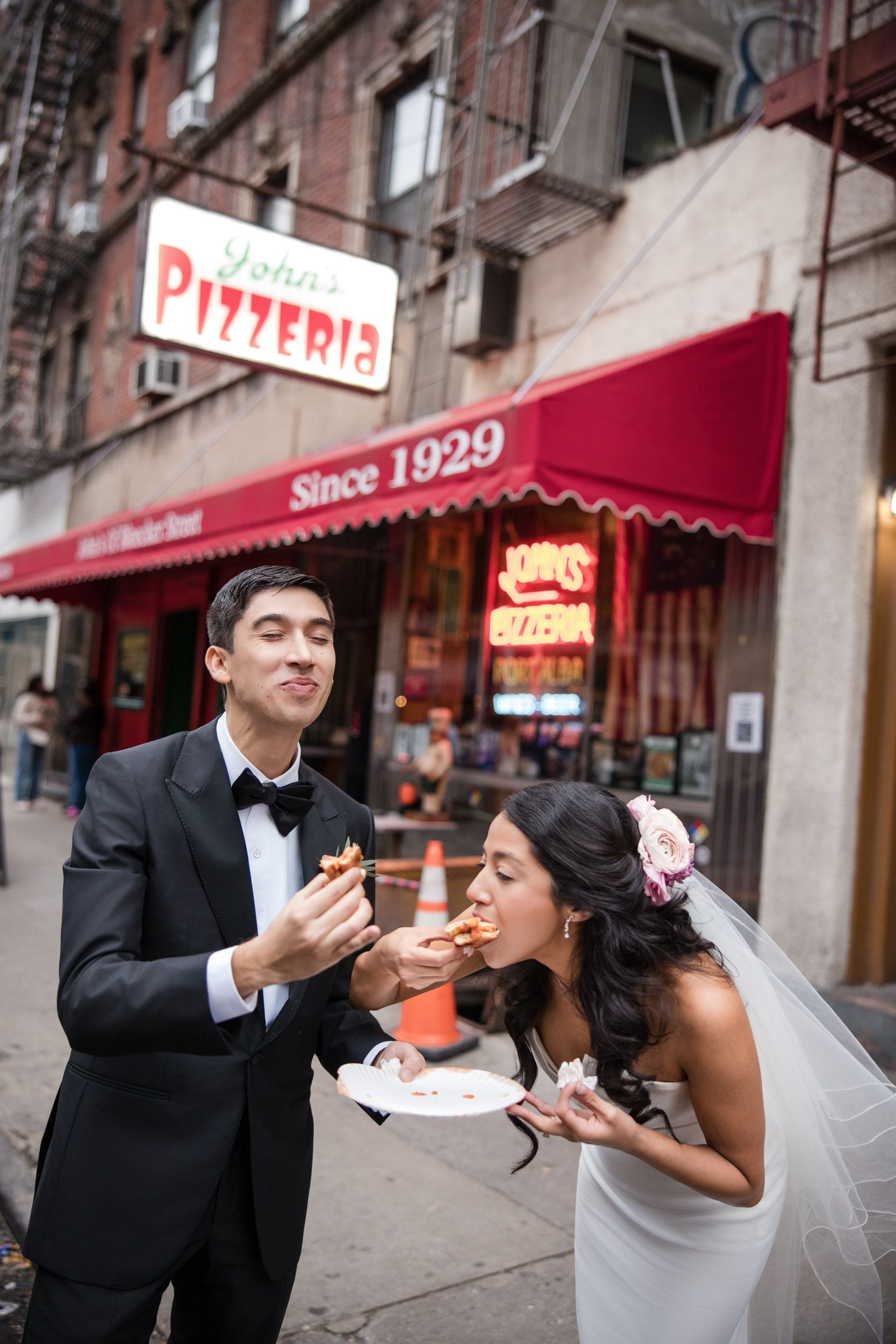 A bride and groom in formal attire eat pizza together outside a pizzeria with a red awning and neon sign, celebrating their St. Patrick’s Cathedral wedding on a lively city sidewalk before heading to Castell Rooftop Lounge.