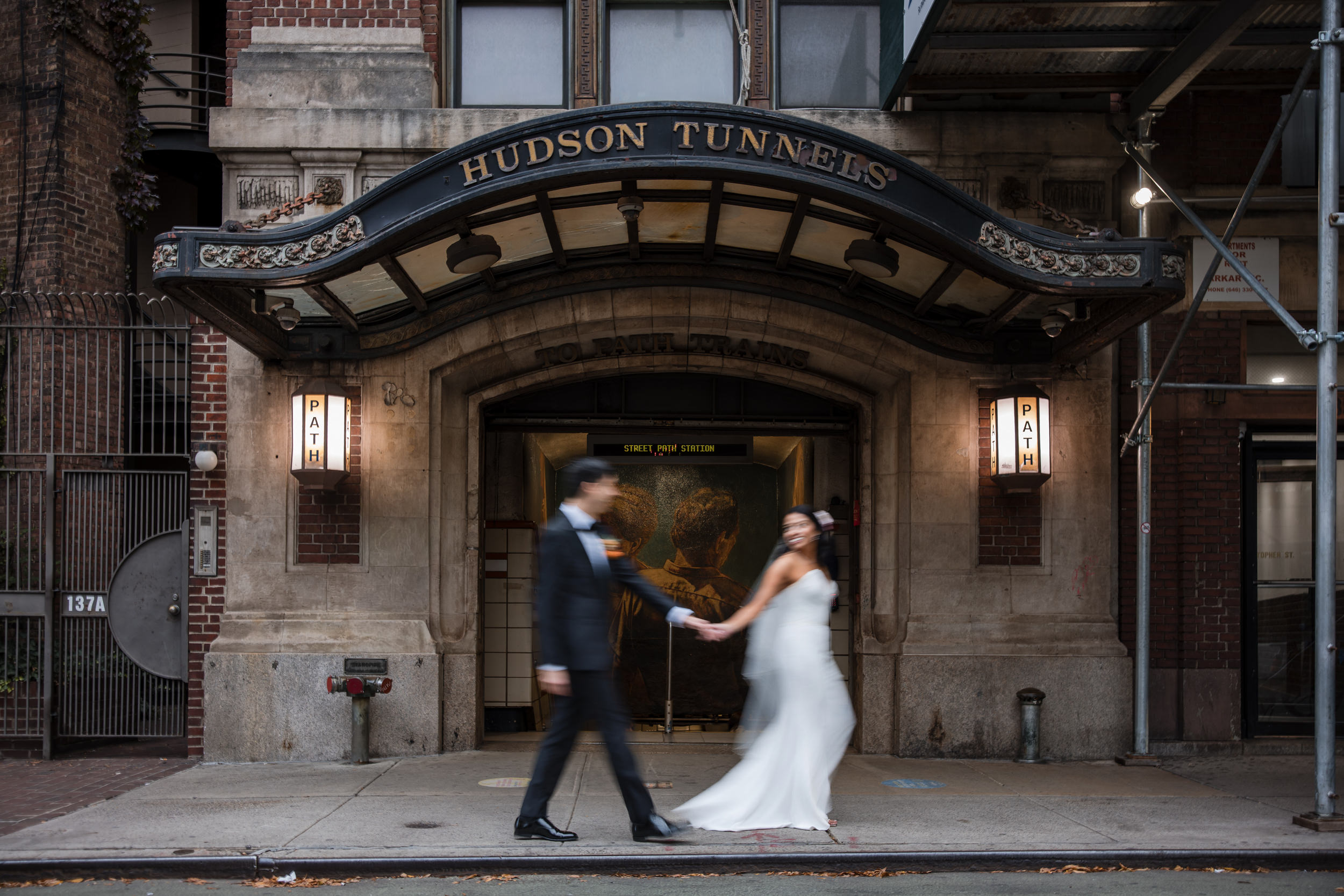 A bride and groom in wedding attire hold hands and walk in front of the Hudson Tunnels entrance, capturing a romantic city moment after their St. Patrick’s Cathedral wedding.