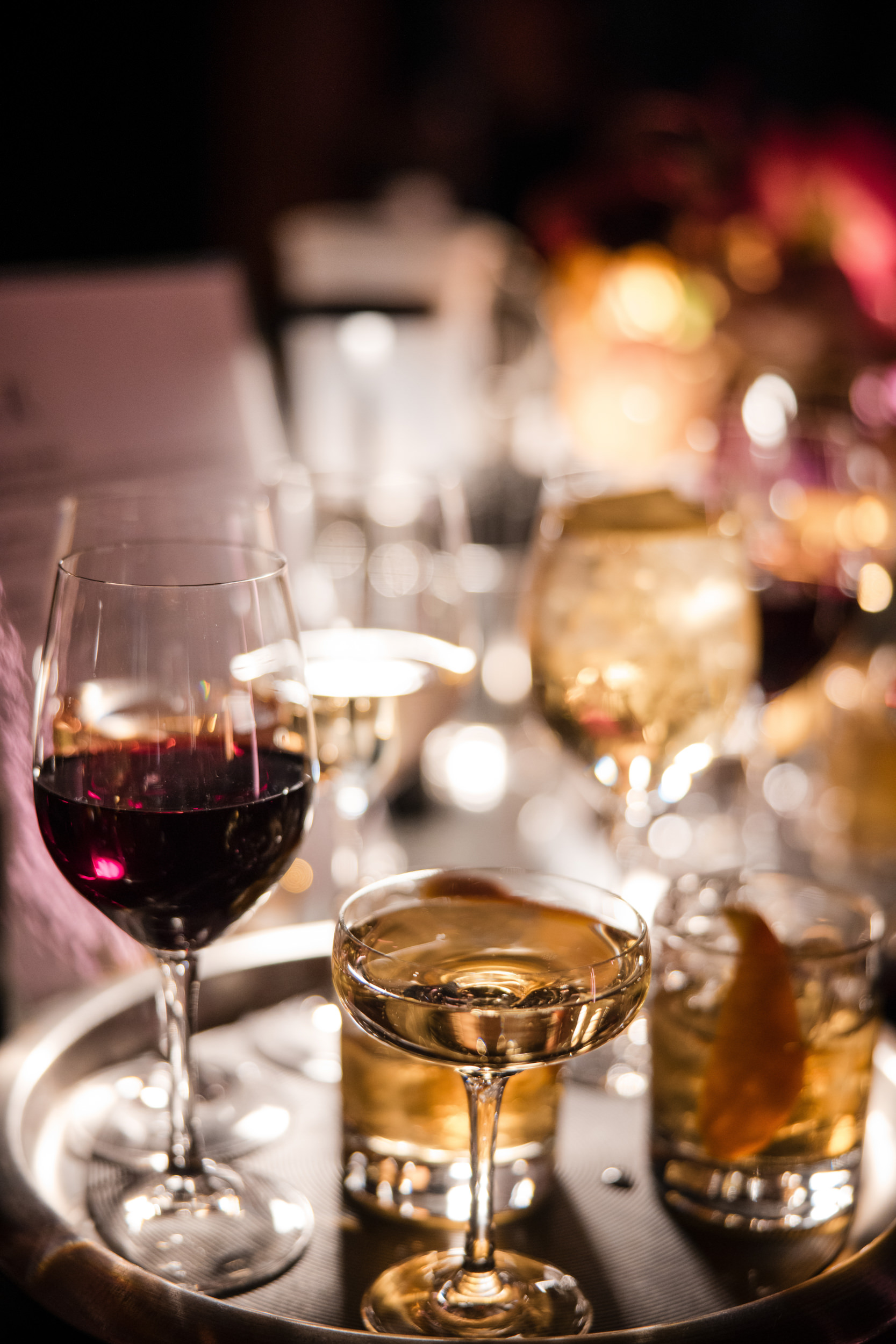 A tray at Castell Rooftop Lounge holds various drinks—a glass of red wine, a coupe cocktail, and more—set on a table with blurred lights in the background, perfect for celebrating after a St. Patrick's Cathedral wedding.