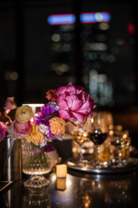 A close-up of a table at Castell Rooftop Lounge, with a floral arrangement, glasses of wine, a lit candle, and blurred city lights—perfect for celebrating after a St. Patrick's Cathedral wedding.