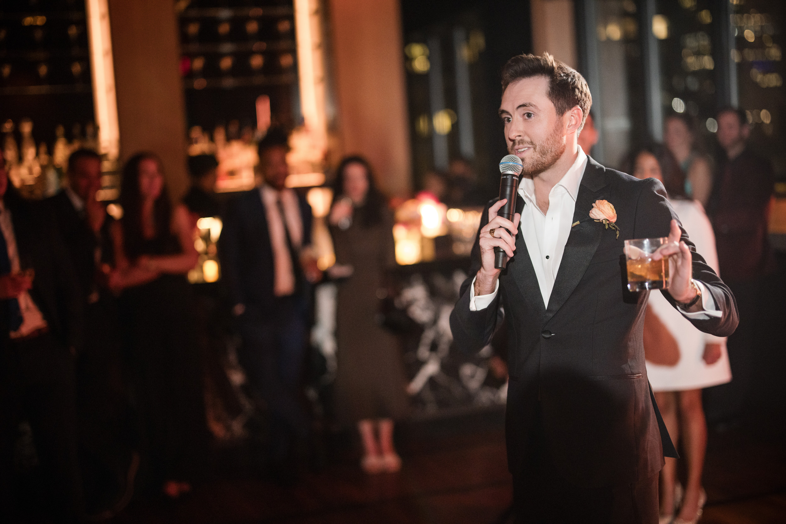 A man in a suit holding a microphone and a drink speaks to an audience at an indoor evening event at Castell Rooftop Lounge; guests, perhaps from a St. Patrick's Cathedral wedding, stand and listen in the background.